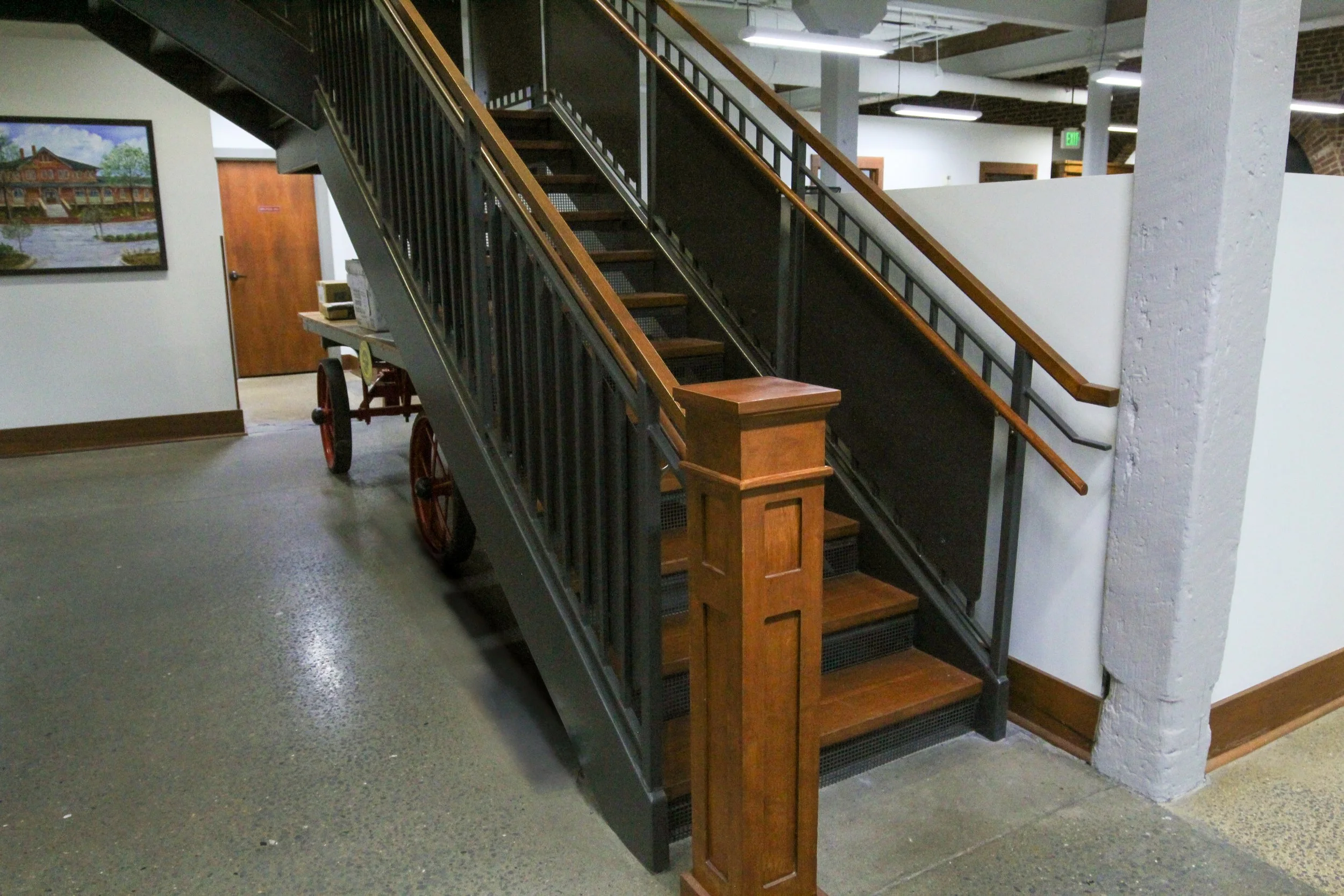 Wooden staircase with black metal railing and handrail, in an interior space with concrete and wood flooring, brick and white walls, and artwork on the wall.