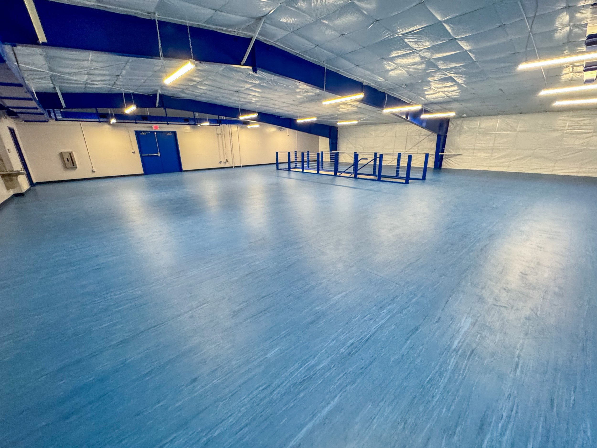 Empty indoor sports or recreational facility with blue flooring, blue walls and a blue door, featuring overhead lighting and a railing near an elevated area.