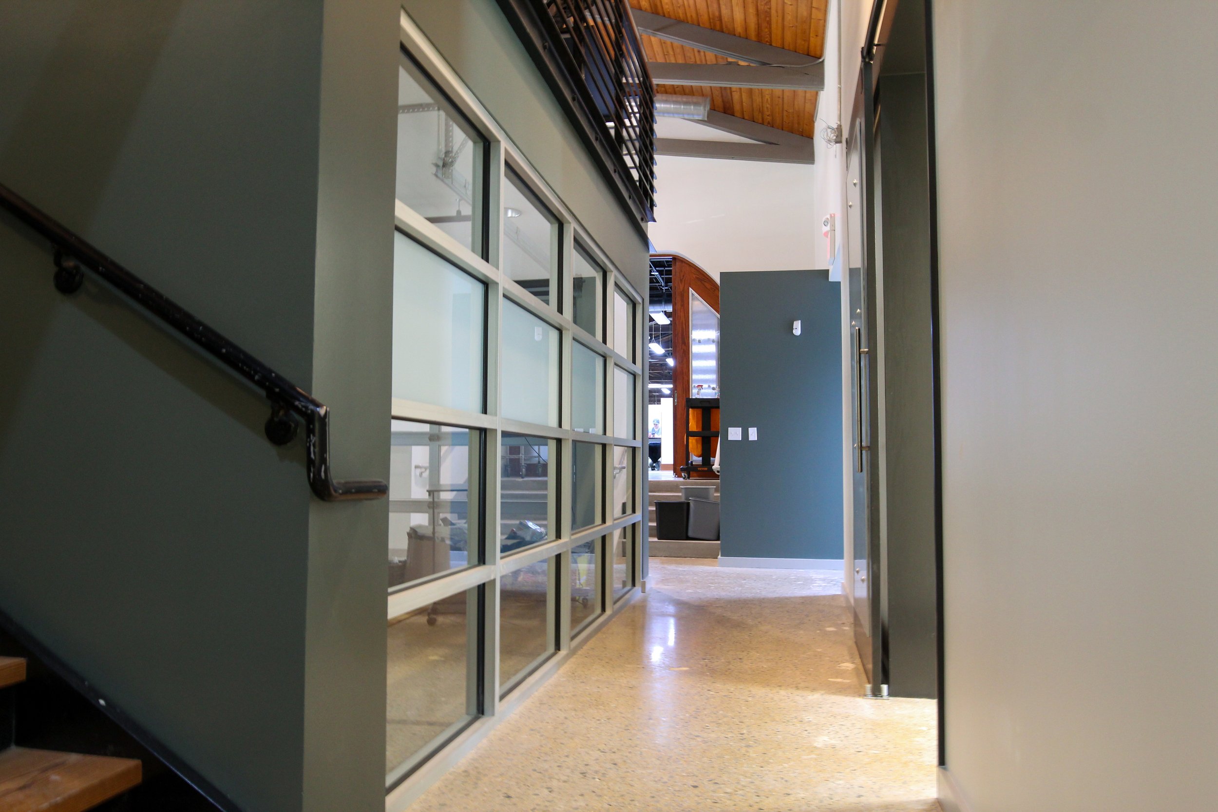 Interior view of an office building aisle, showing stairs on the left, modern glass partitions on the left, a partial wall with a hook, and an open space with some furniture and a wooden ceiling in the background.
