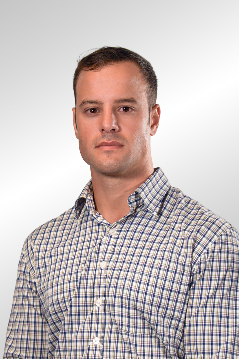 A young man with short brown hair, wearing a checked button-up shirt, standing against a plain light background.