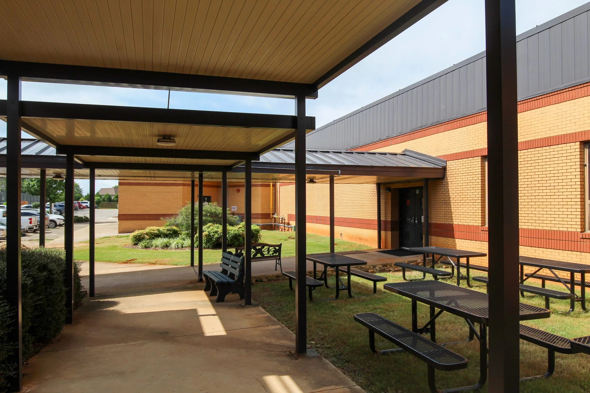 View of a school or institutional building exterior with a covered outdoor area, benches, and lush green landscaping.