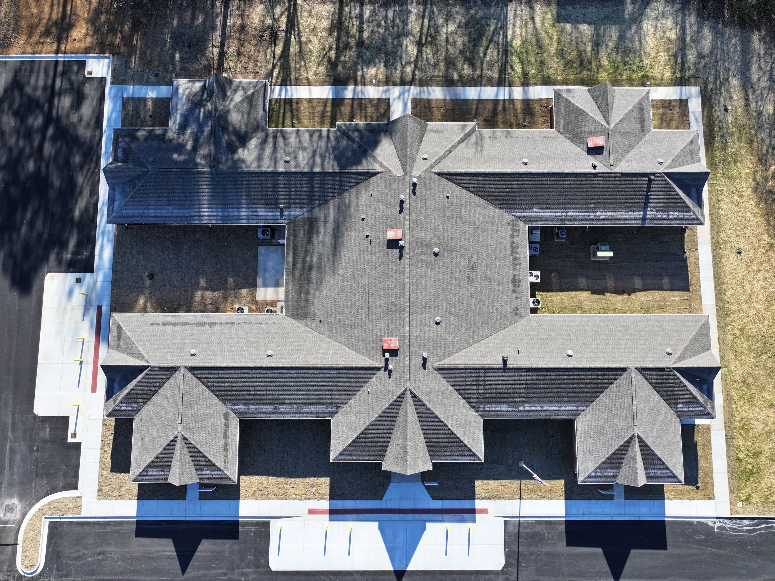 An aerial view of a building with multiple roof sections, surrounded by sidewalks and parking areas, with some trees and ground visible around the building.