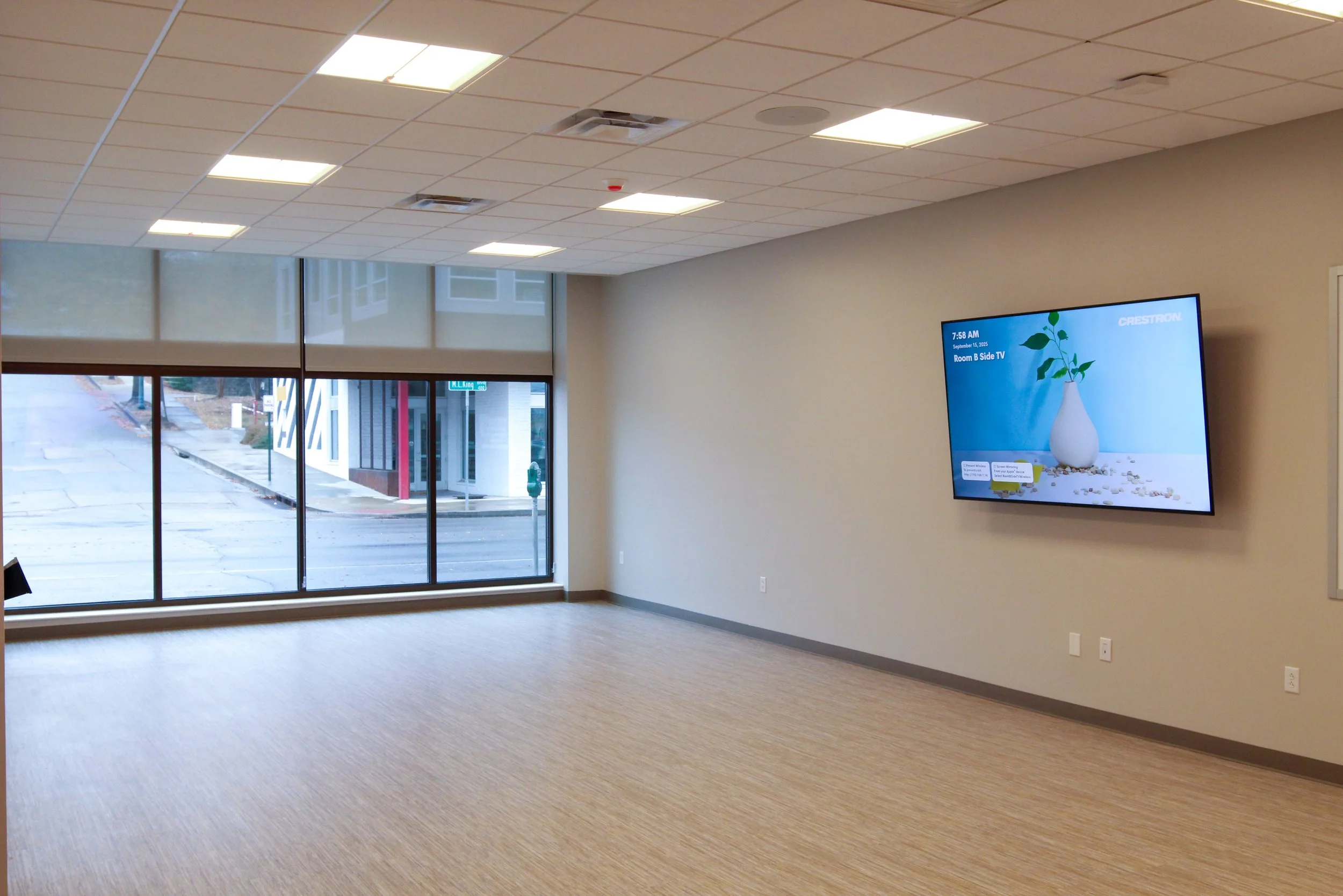 Empty conference or meeting room with large front window, beige carpet, and wall-mounted television displaying a blue background with a white vase and green plant.