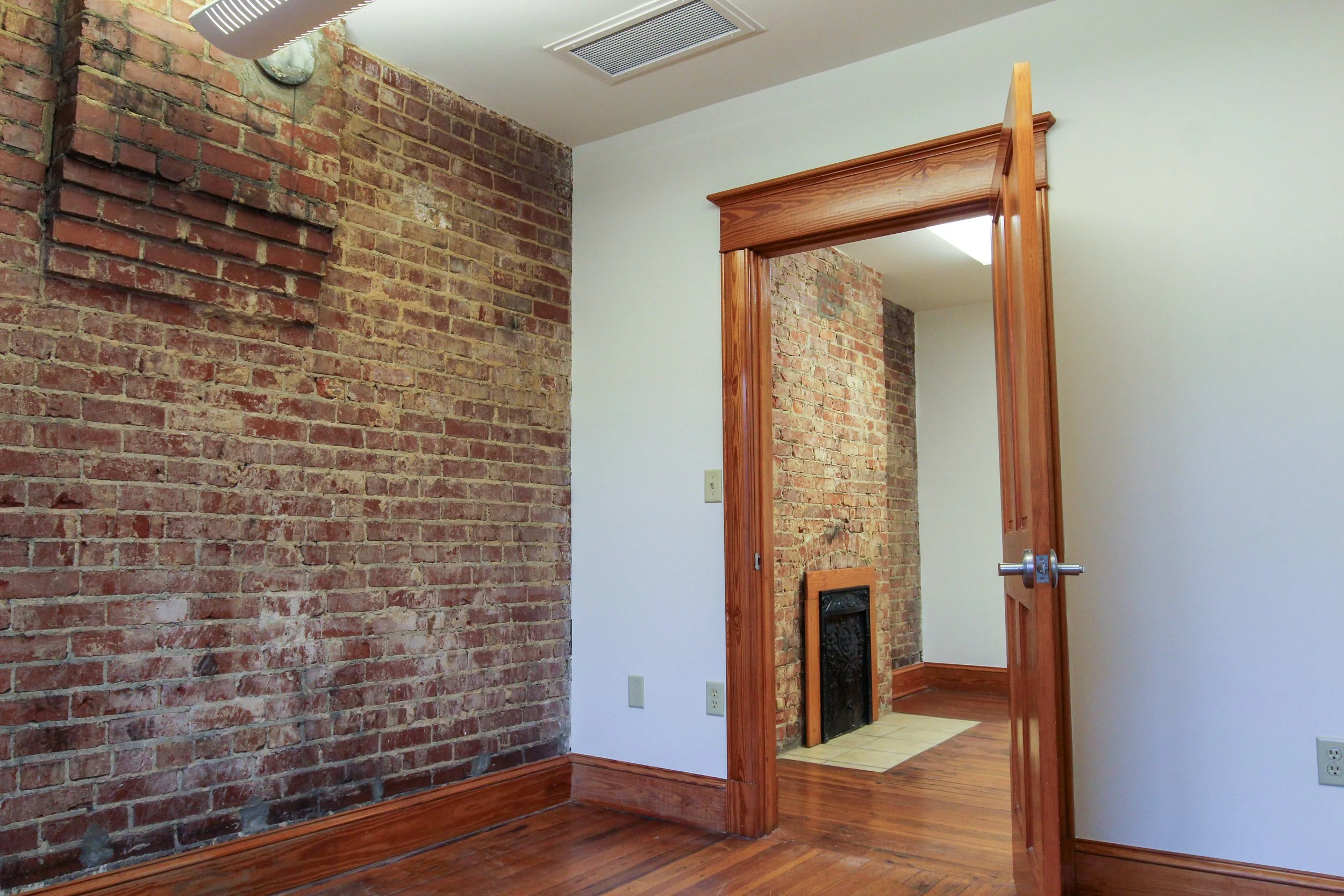 Interior room with exposed red brick wall, hardwood floor, wooden door frame, and a doorway leading to another room with brick wall and fireplace