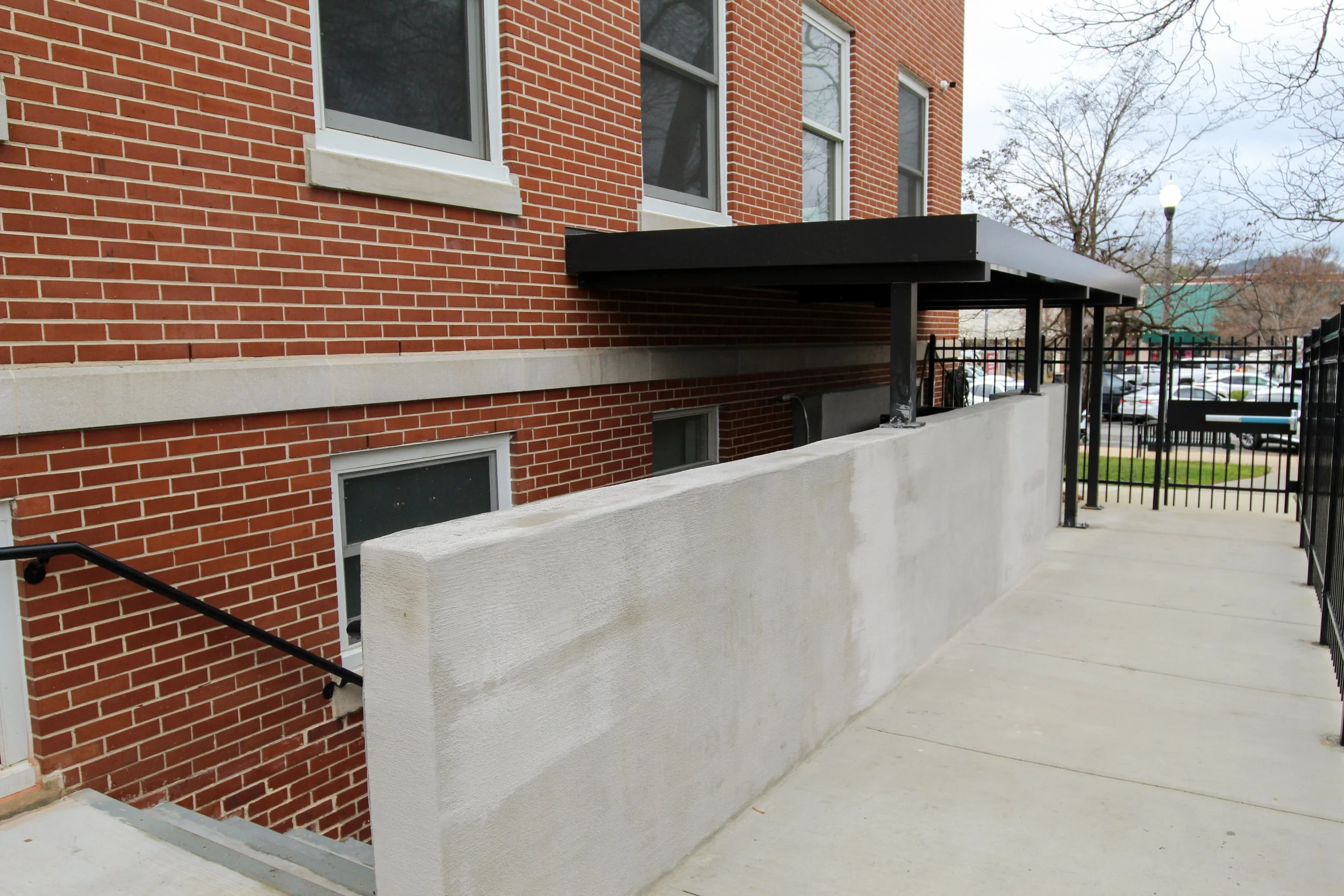 Exterior view of a brick building with a concrete walkway, black handrail, and small concrete wall. A black awning extends over part of the walkway, with a fenced parking lot in the background.