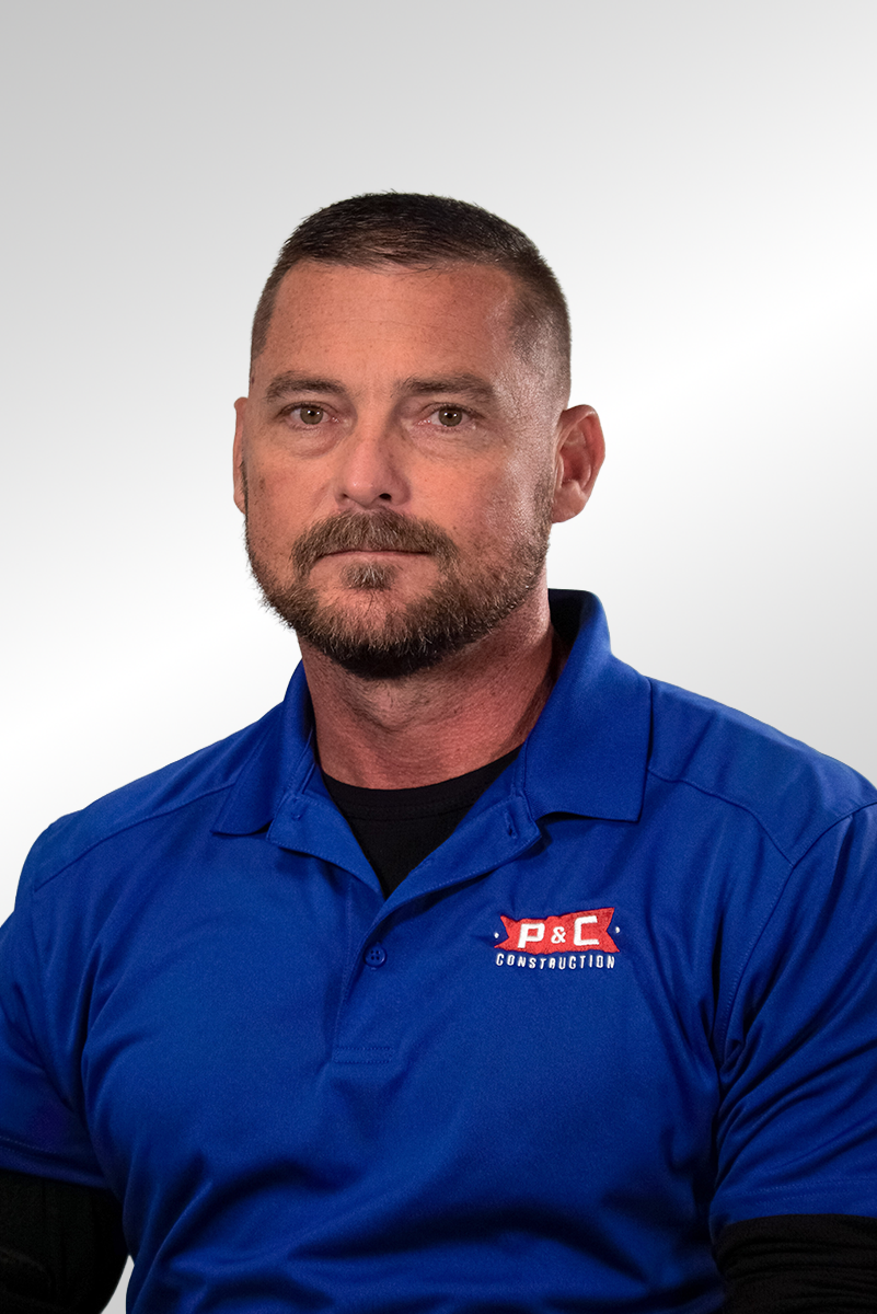 A man with short brown hair, a beard, and a light complexion wearing a black and gray patterned polo shirt, sitting against a plain dark background.