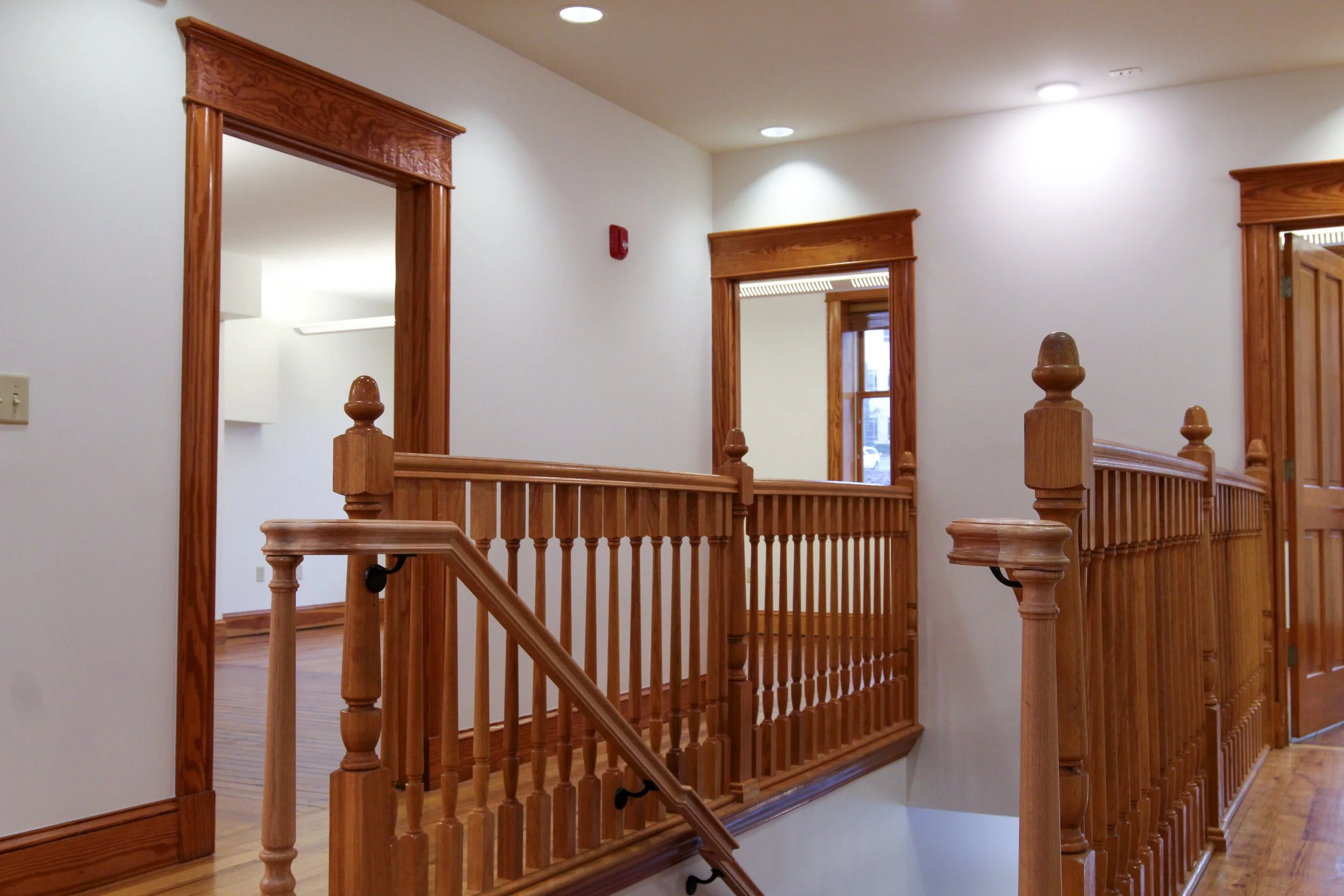 Interior of a house with wooden stair railing, door frames, and hardwood floors, with white walls and ceiling lights.