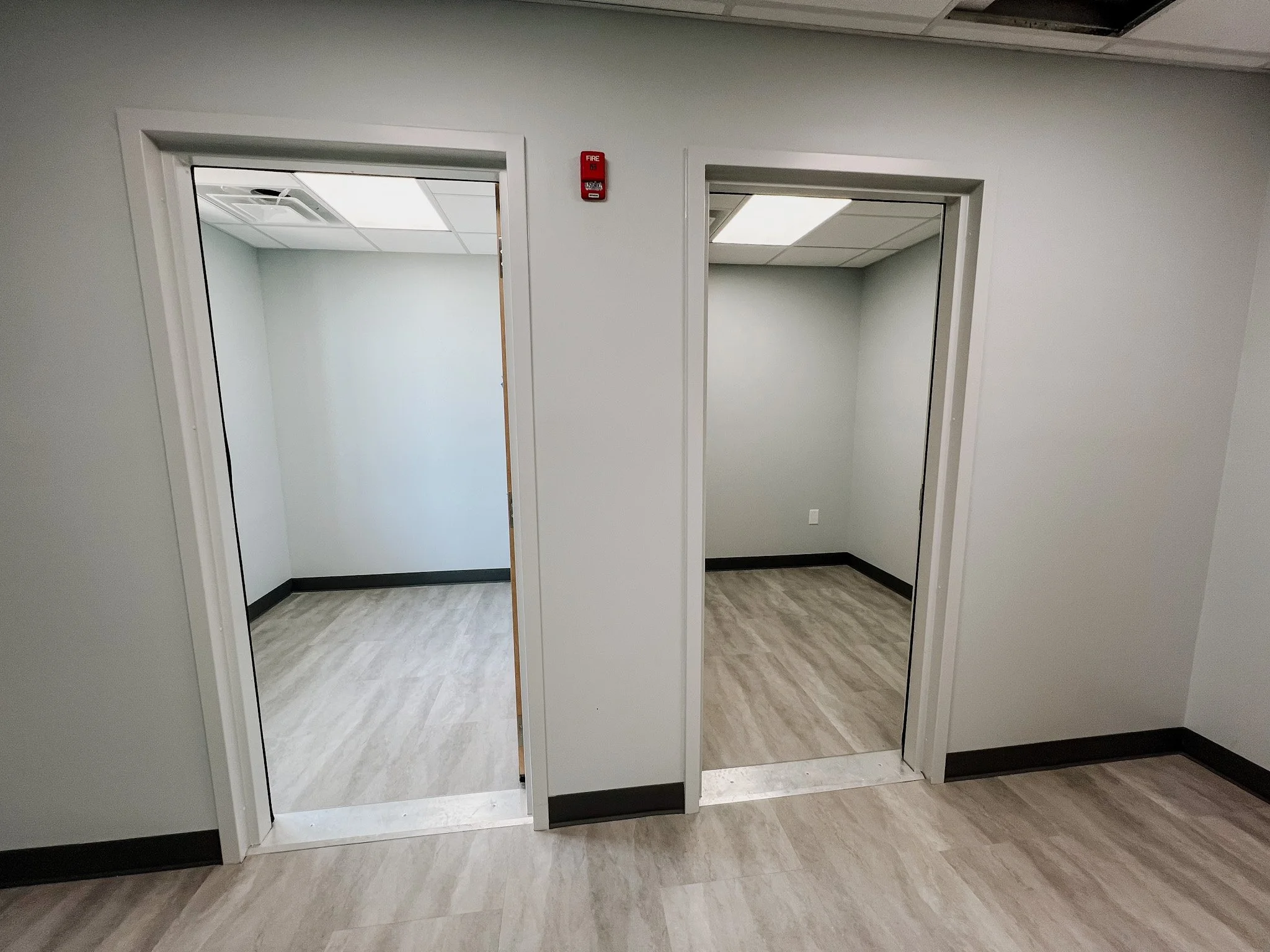 Two empty rooms with gray walls, wooden flooring, black baseboards, and open doorways, viewed from the hallway.
