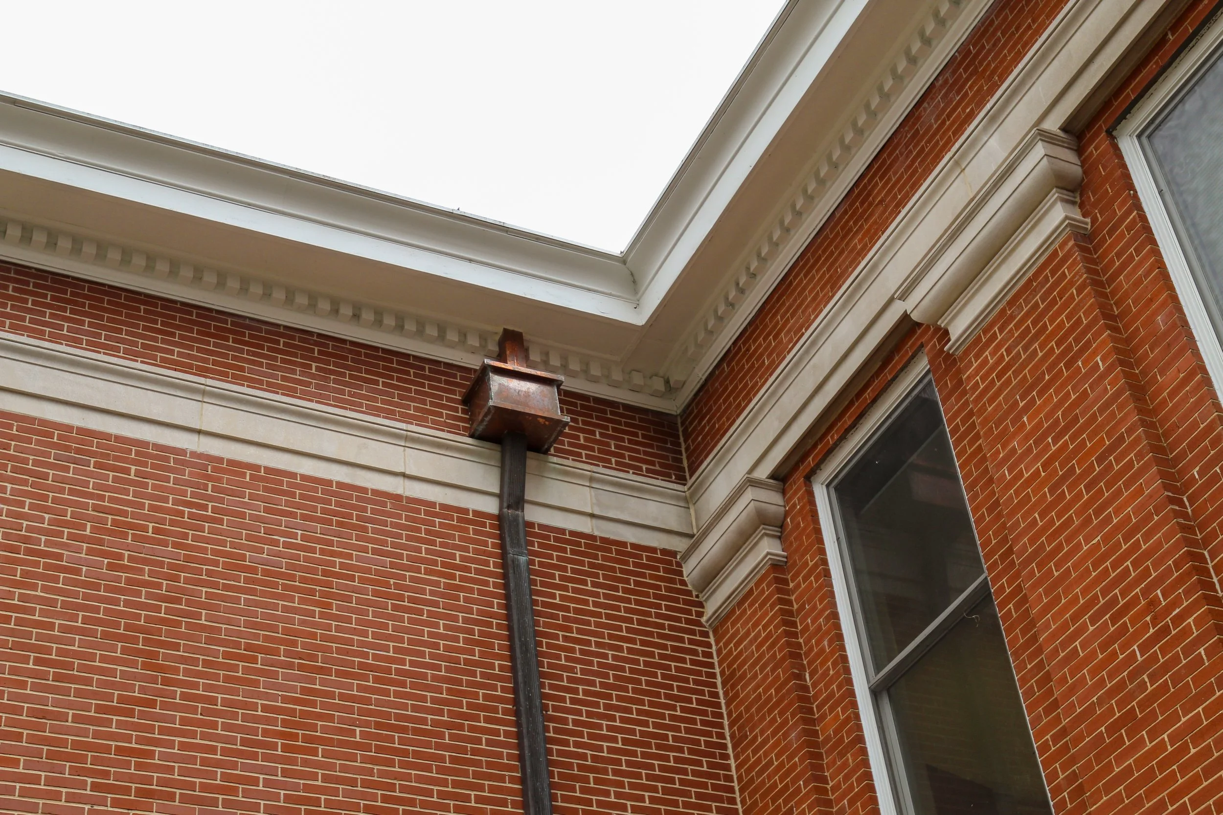 Close-up of a rusty metal ventilation box connected to a downspout pipe on the corner of a brick building with decorative moldings and windows.