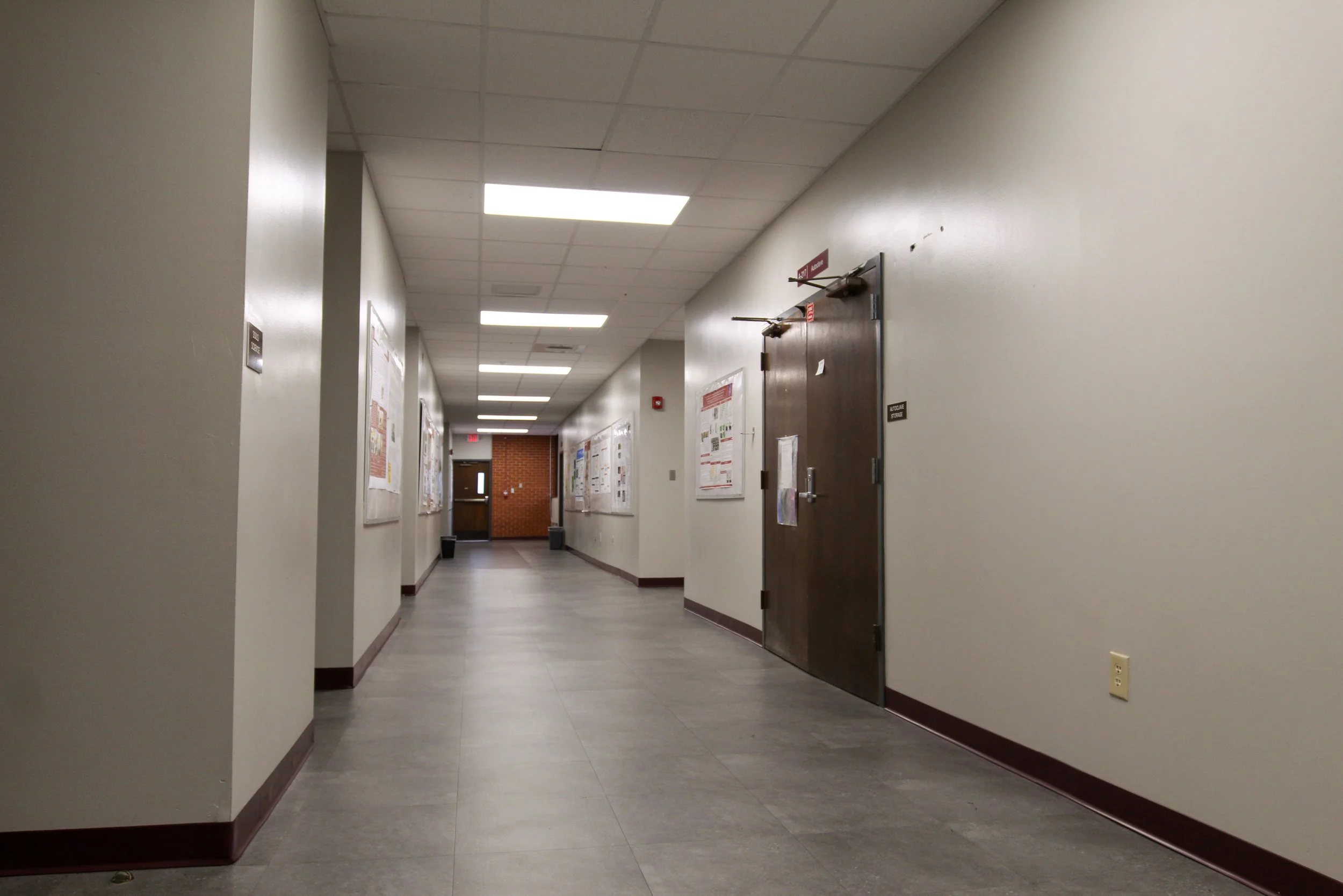 Empty hallway in a building with cream walls, gray tile floor, hanging bulletin boards, and a dark wooden door on the right.
