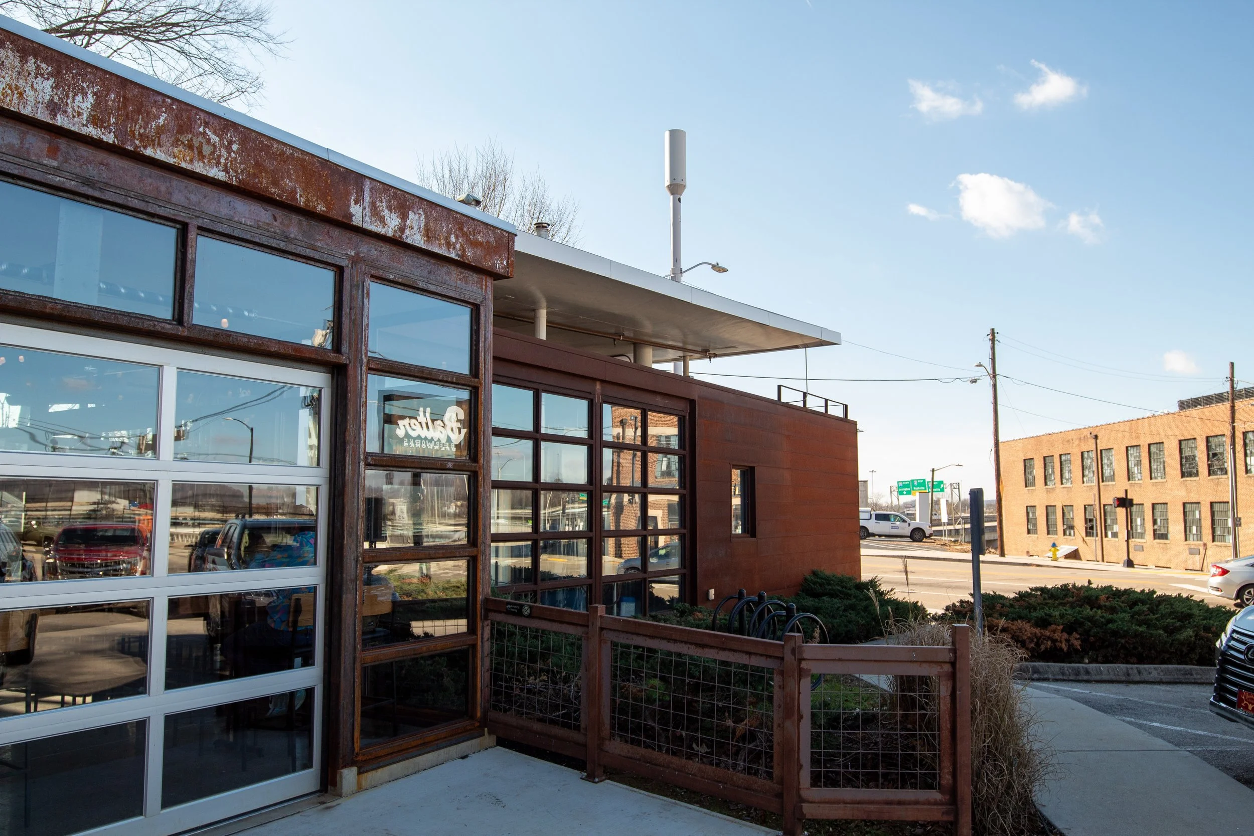 Exterior view of a modern building with large glass windows and rust-colored walls, situated on a city street with cars and another brick building in the background.