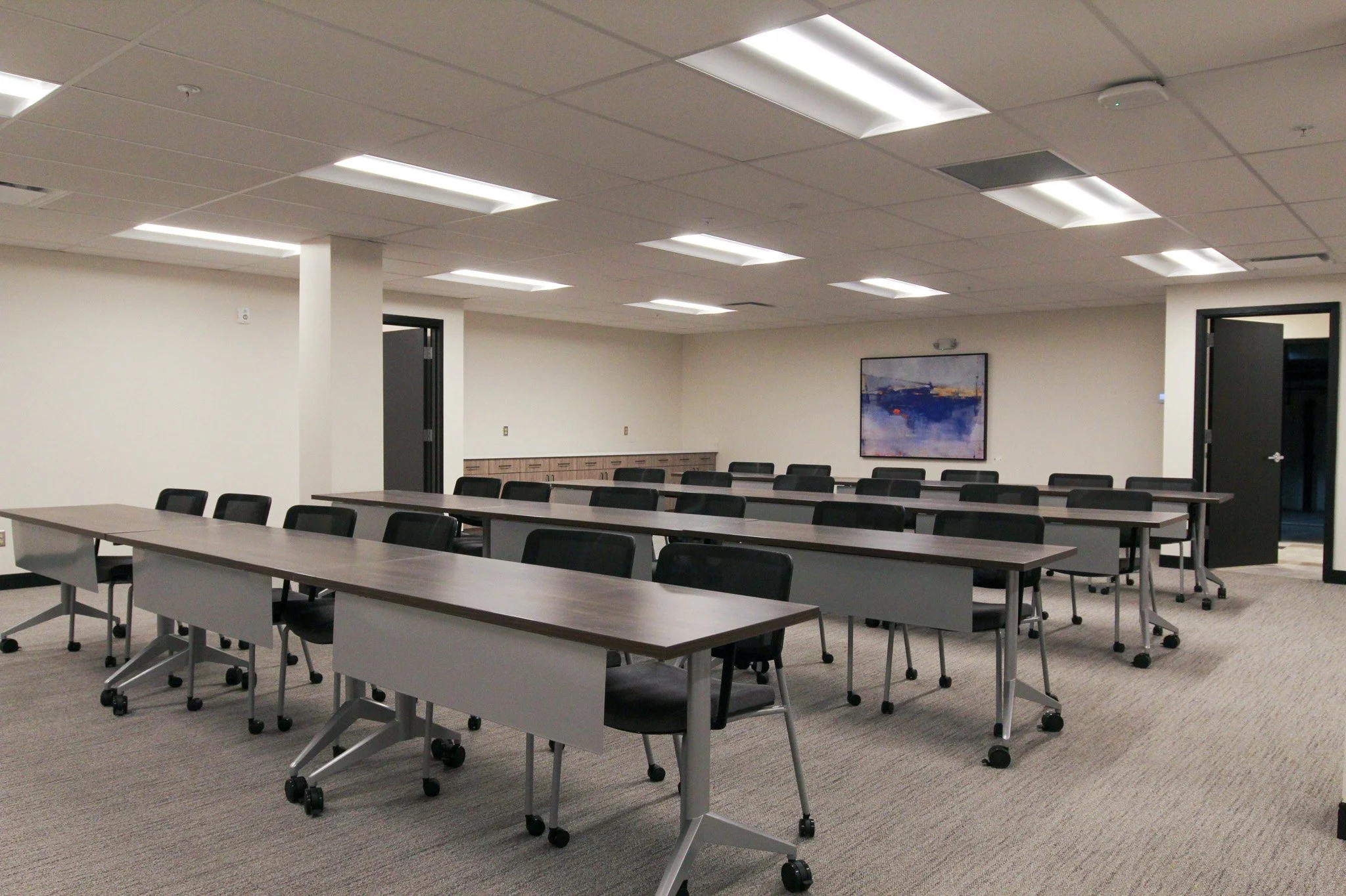 Empty conference room with rows of tables and chairs, white walls, ceiling lights, a framed landscape painting, and a door.
