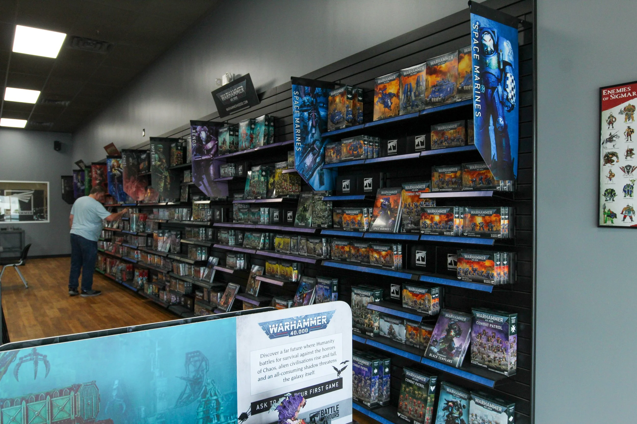 A man inspecting a display of Warhammer 40,000 tabletop game boxes on shelves inside a game or hobby store. The shelves hold various Warhammer 40,000 boxes featuring models and expansions. There is a sign advertising the game and a poster with charac
