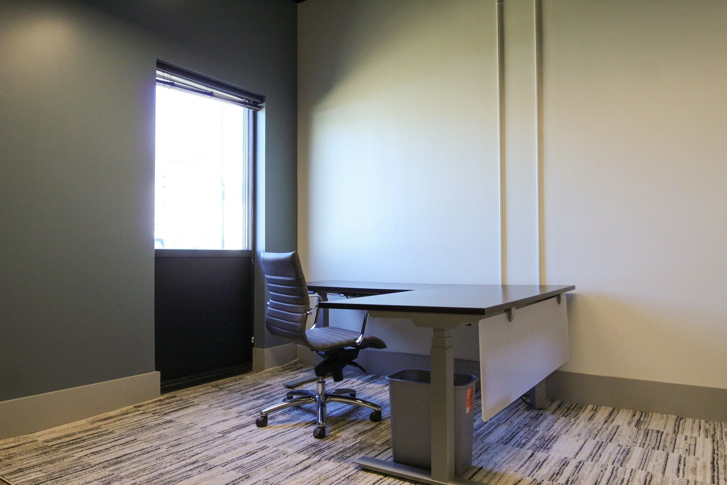 Empty office with a desk, office chair, window with blinds, and carpeted floor.