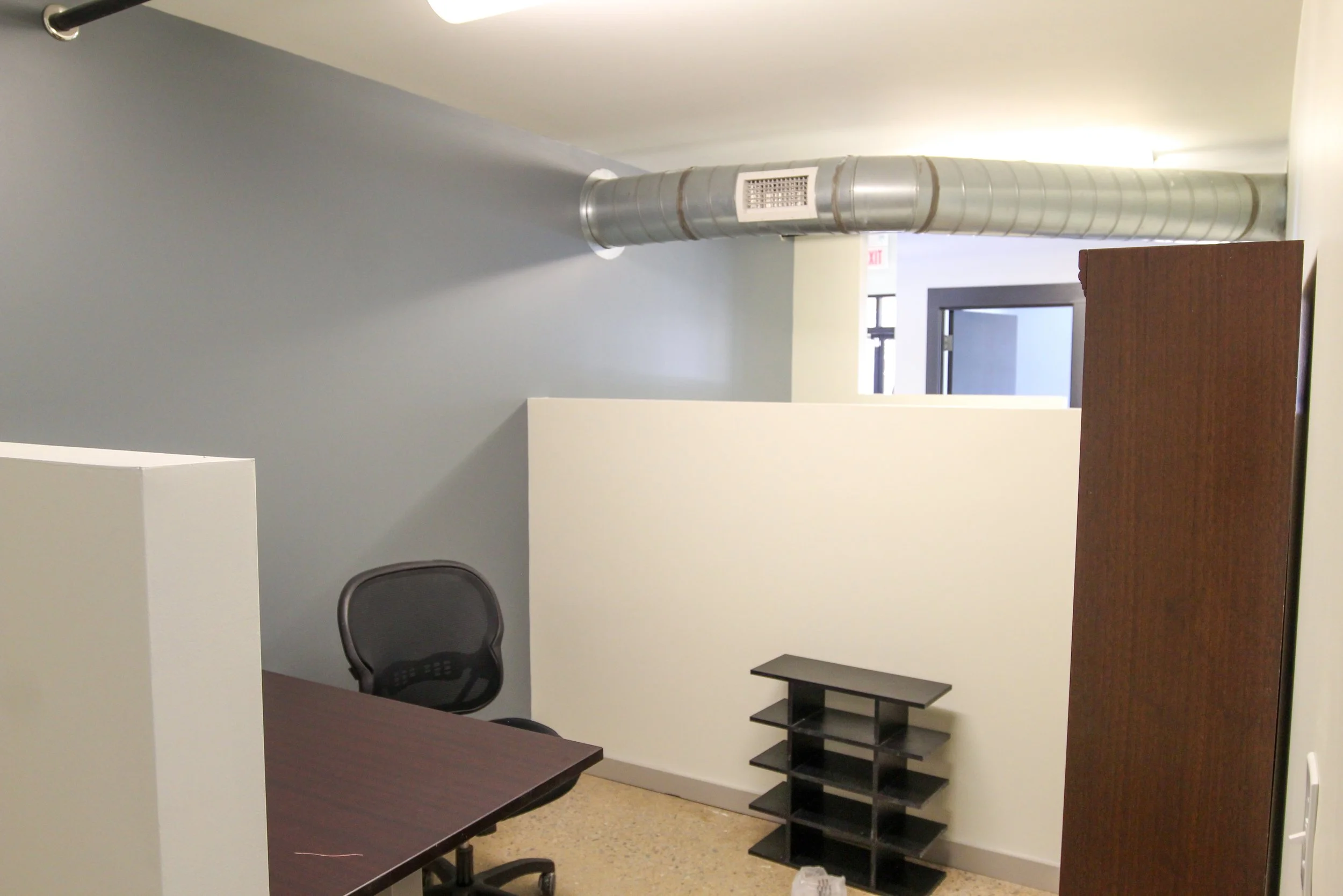 An office cubicle with a black swivel chair, a brown desk, and a black shelving unit. There is a white partition wall and a gray wall behind it, with an exposed ventilation duct on the ceiling.