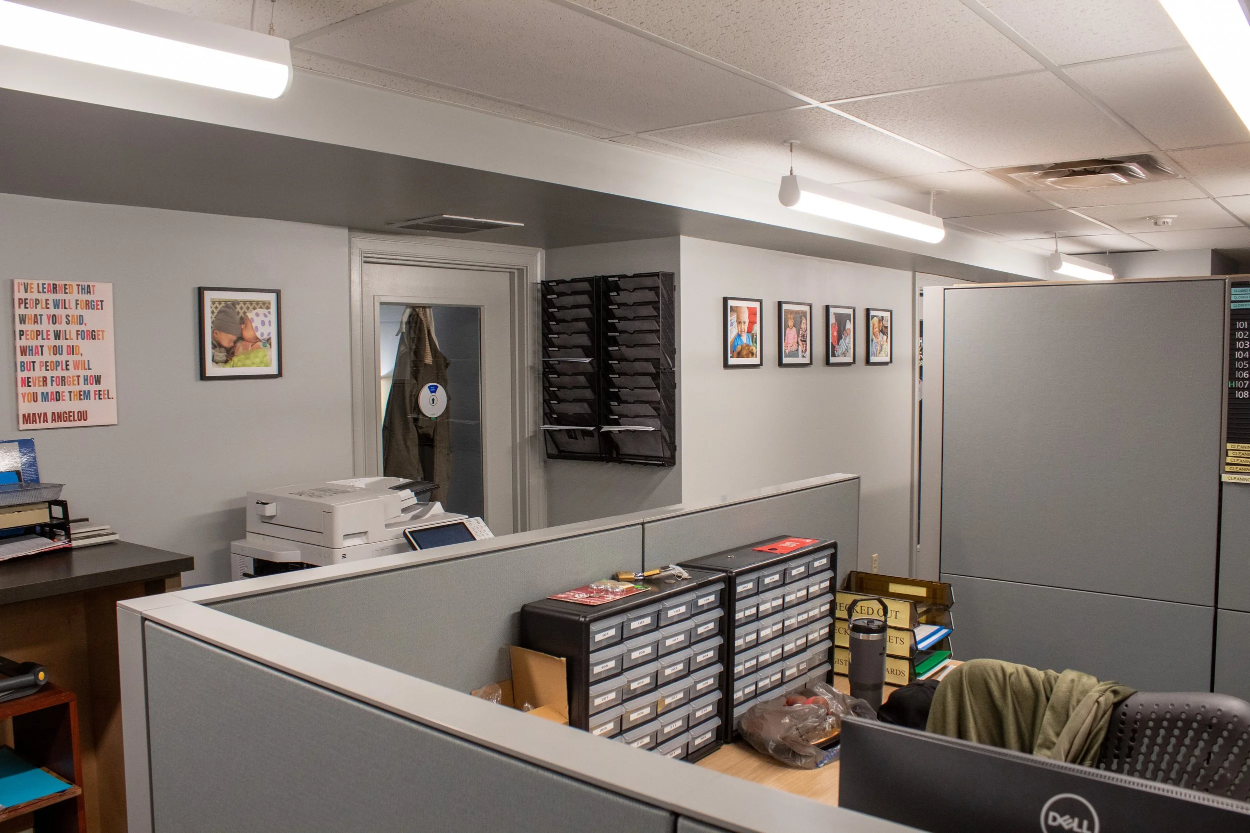 Office cubicles with filing drawers, a desk with computer and supplies, framed pictures on the wall, a printer, and various office items.