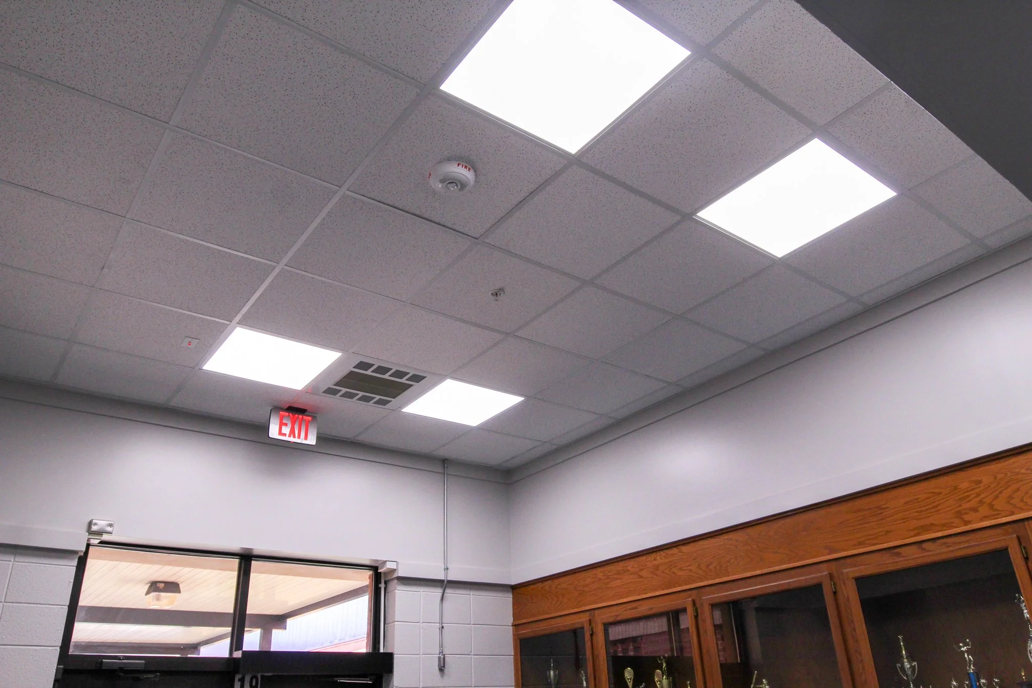 Ceiling of an indoor room with fluorescent light panels, smoke detector, fire alarm, and an exit sign. Part of a wooden display cabinet and glass trophies are visible below.
