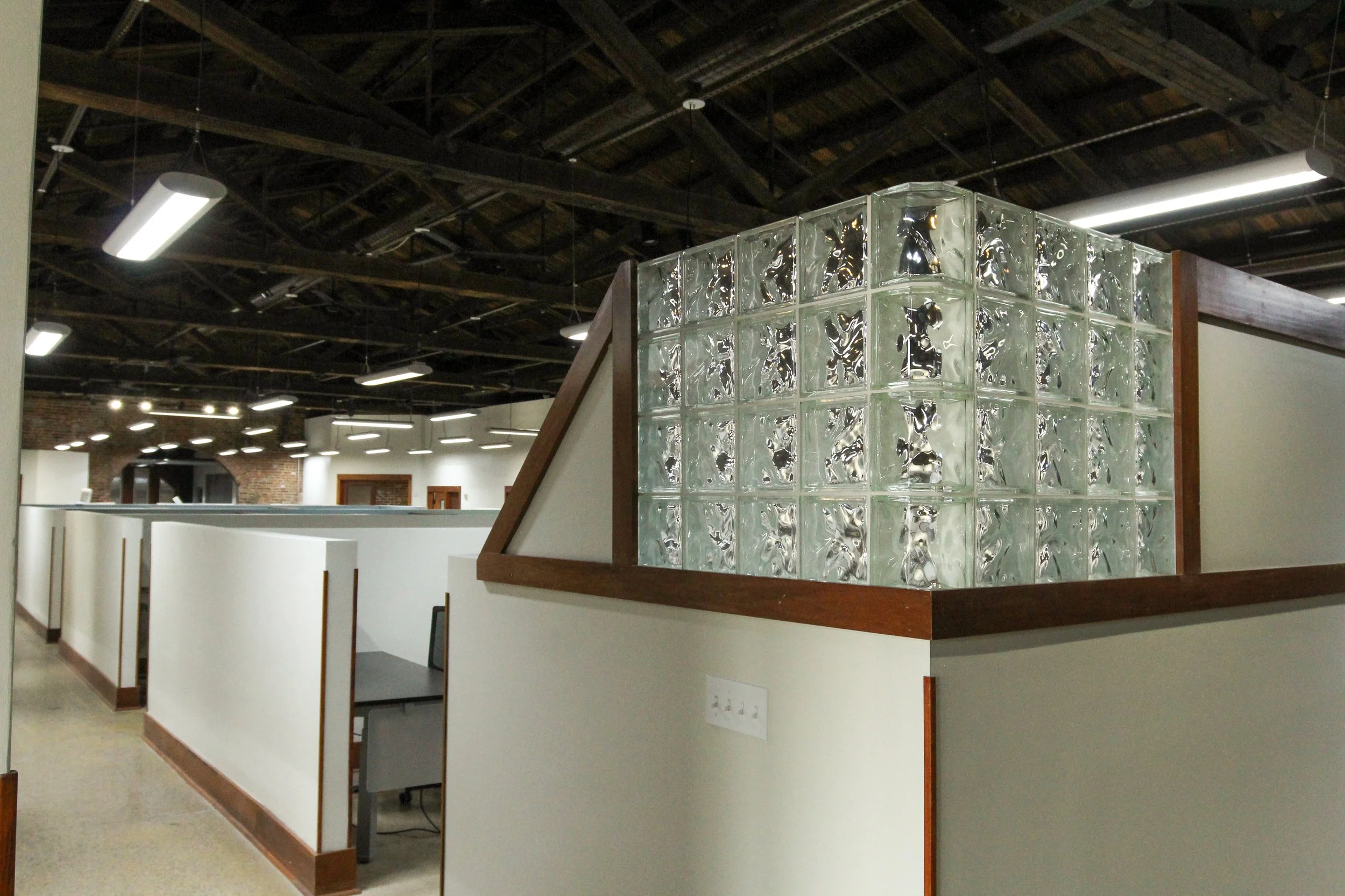Interior view of an office space with cubicles, a glass block wall, and exposed ceiling beams with industrial lighting.