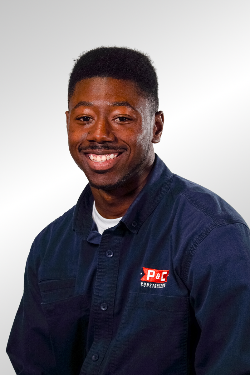 A young man with short, curly hair and a beard, wearing a blue polo shirt, looking directly at the camera with a neutral expression, against a solid gray background.