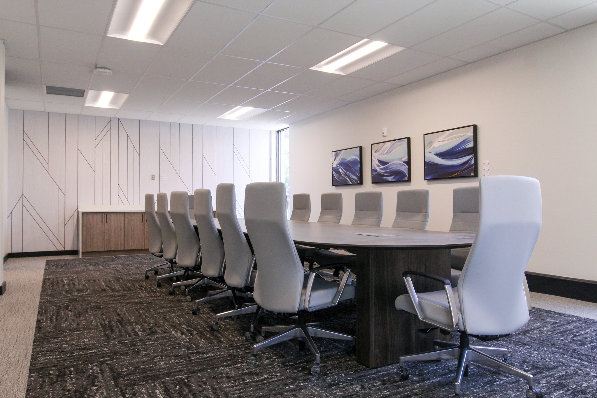 A modern conference room of Simmons Bank in Athens, TN with a long oval table, white swivel chairs, three framed abstract art pieces on the wall, a large window, and a patterned carpet.