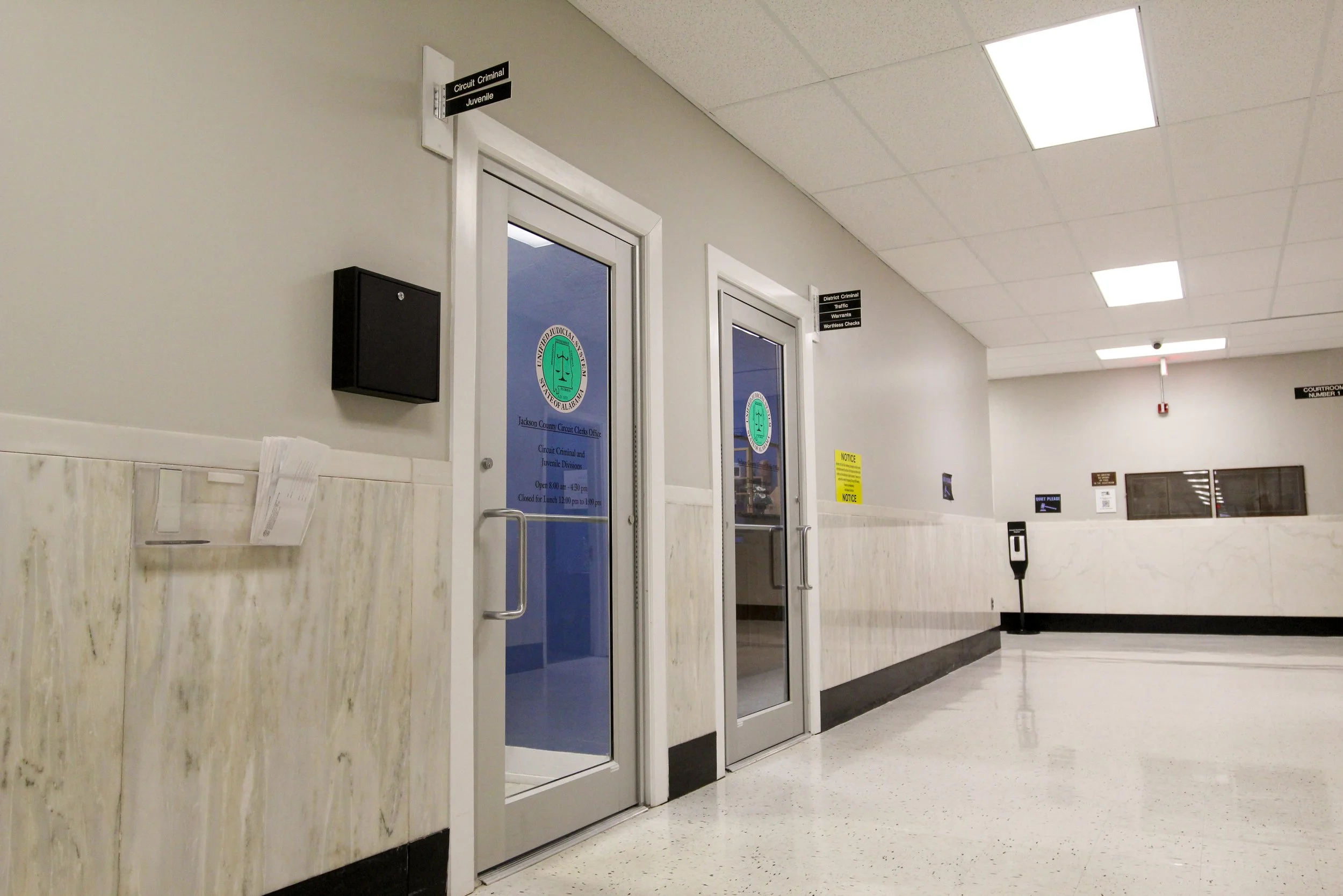 Empty courthouse corridor with two glass doors labeled 'Circuit Criminal Juvenile' and various signs and notice boards on the wall.