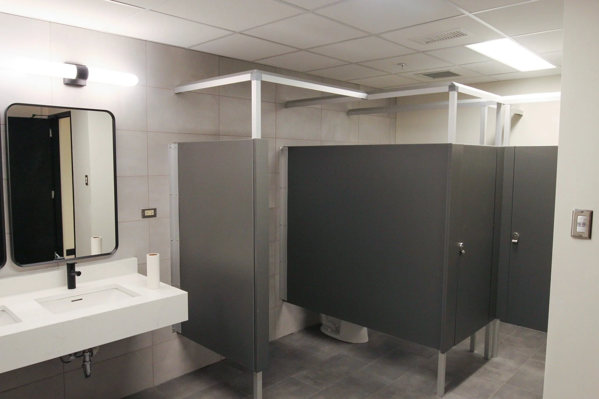 A women's public bathroom with gray stall partitions, a white sink with a black faucet, a mirror, and a large fluorescent light fixture on the ceiling.