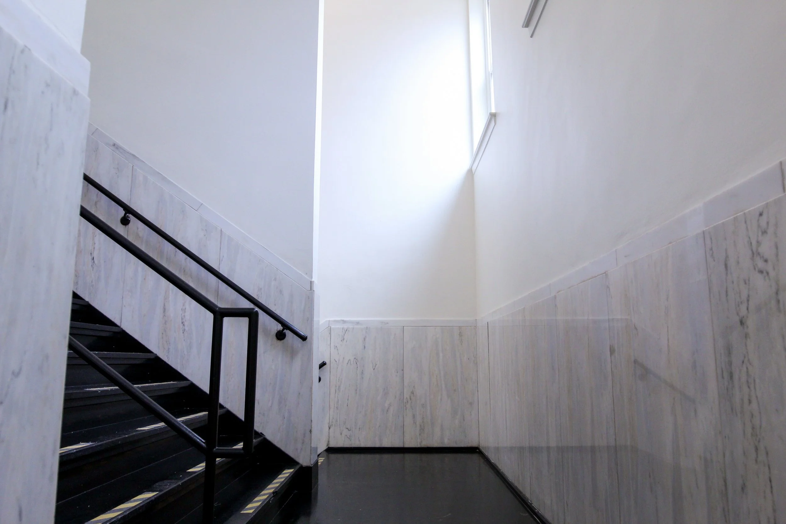 Interior view of a staircase with white marble walls, black steps, and a black metal handrail, illuminated by natural light from a high window.