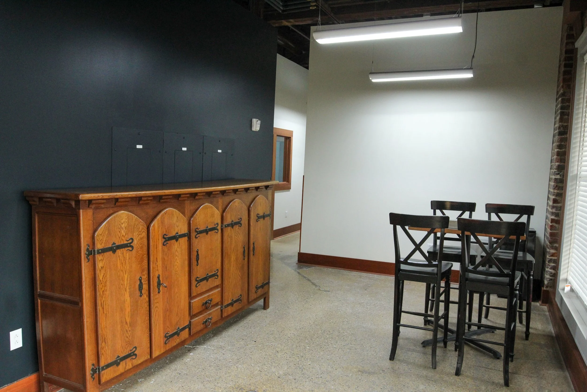 Interior of a room with a dark accent wall, wooden cabinet, and a black table with four chairs near a large window with blinds.