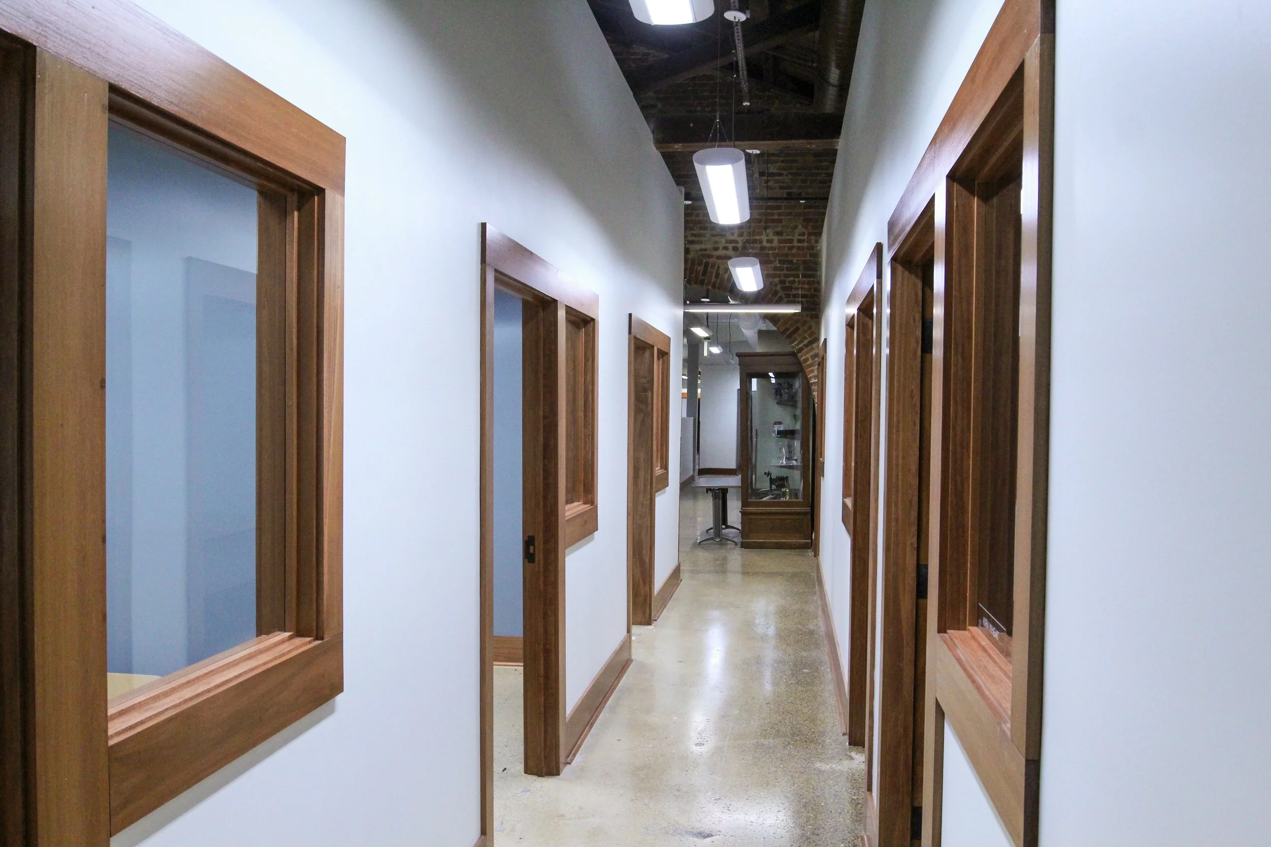 Interior view of a hallway with white walls, wooden trim around windows and doors, and exposed brick ceiling with hanging lights.