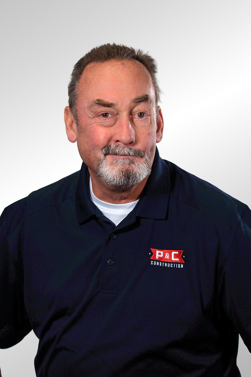 A middle-aged man with gray hair and a beard, wearing a black polo shirt, smiling slightly with one eye closed against a plain dark background.
