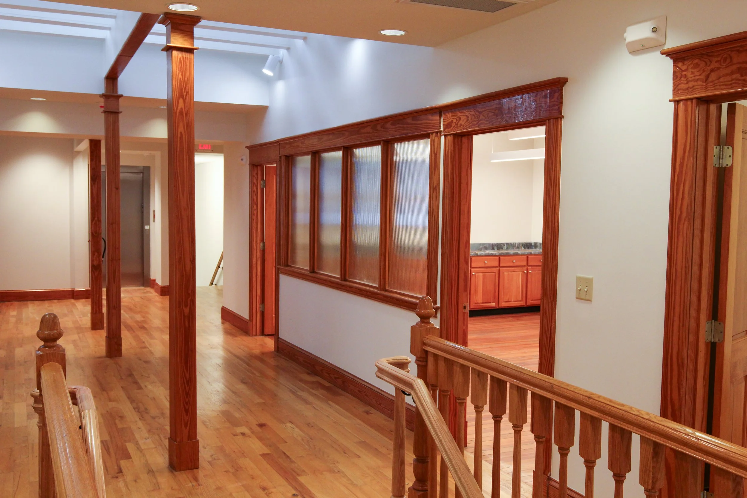 Interior view of a room with wooden trim and flooring, including a corridor, a partial view of a kitchen with wooden cabinets and a granite countertop visible through an open doorway, and large windows with frosted glass panels