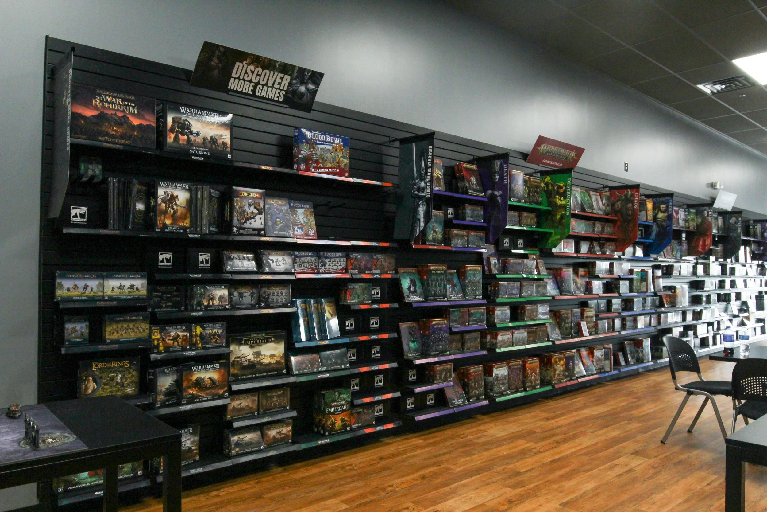 Video game store shelf displaying tabletop miniature game boxes, including Warhammer, Blood Bowl, and Lord of the Rings, with a sign that reads 'Discover More Games'. Remodeled by P&C Construction, Inc. - Chattanooga, Tn.