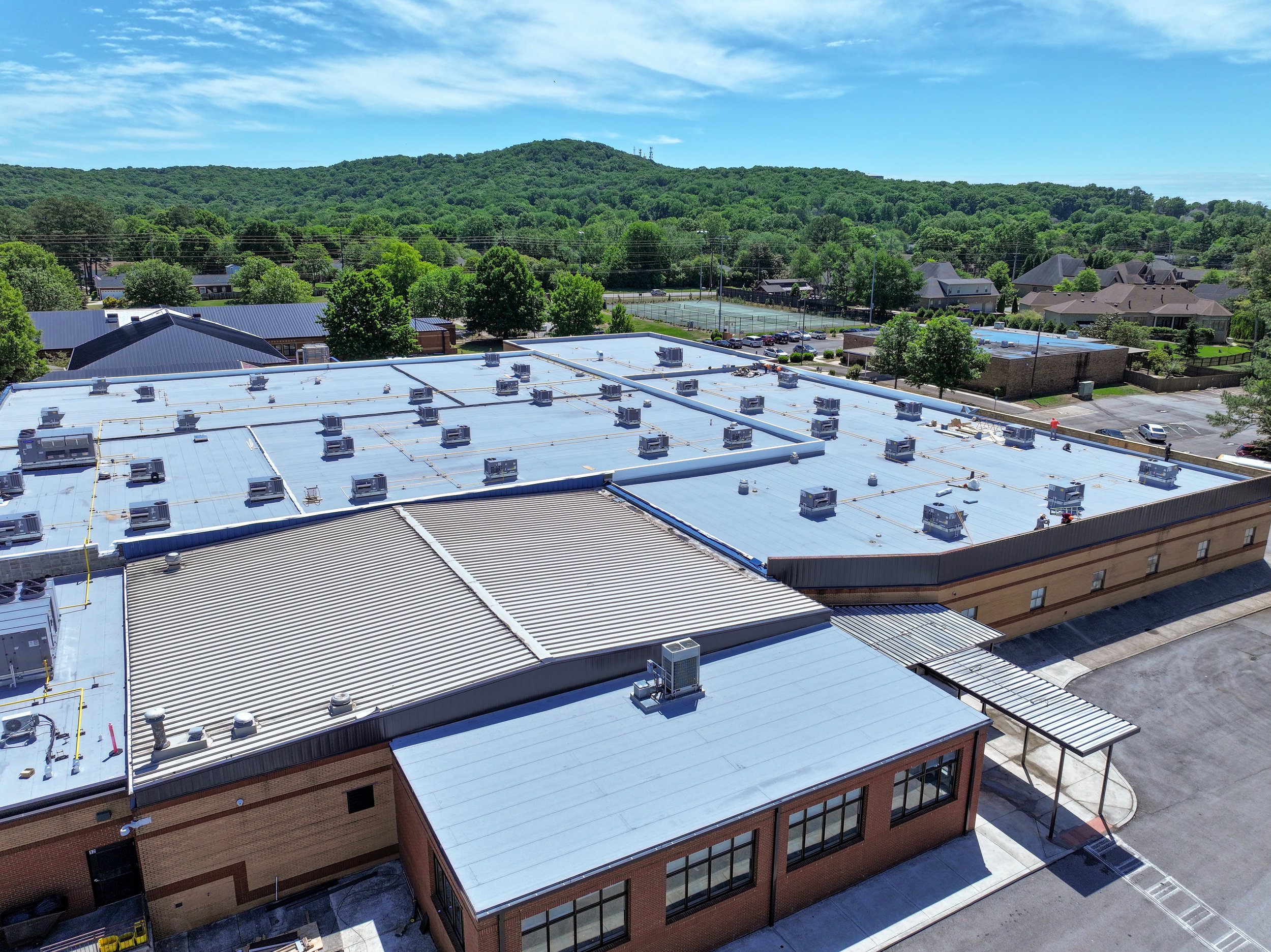 View of a building rooftop with HVAC units, overlooking a parking lot, tennis courts, residential houses, and a hill covered with trees in the background under a blue sky with clouds. Remodeled by P&C Construction, Inc. - Chattanooga, Tn.