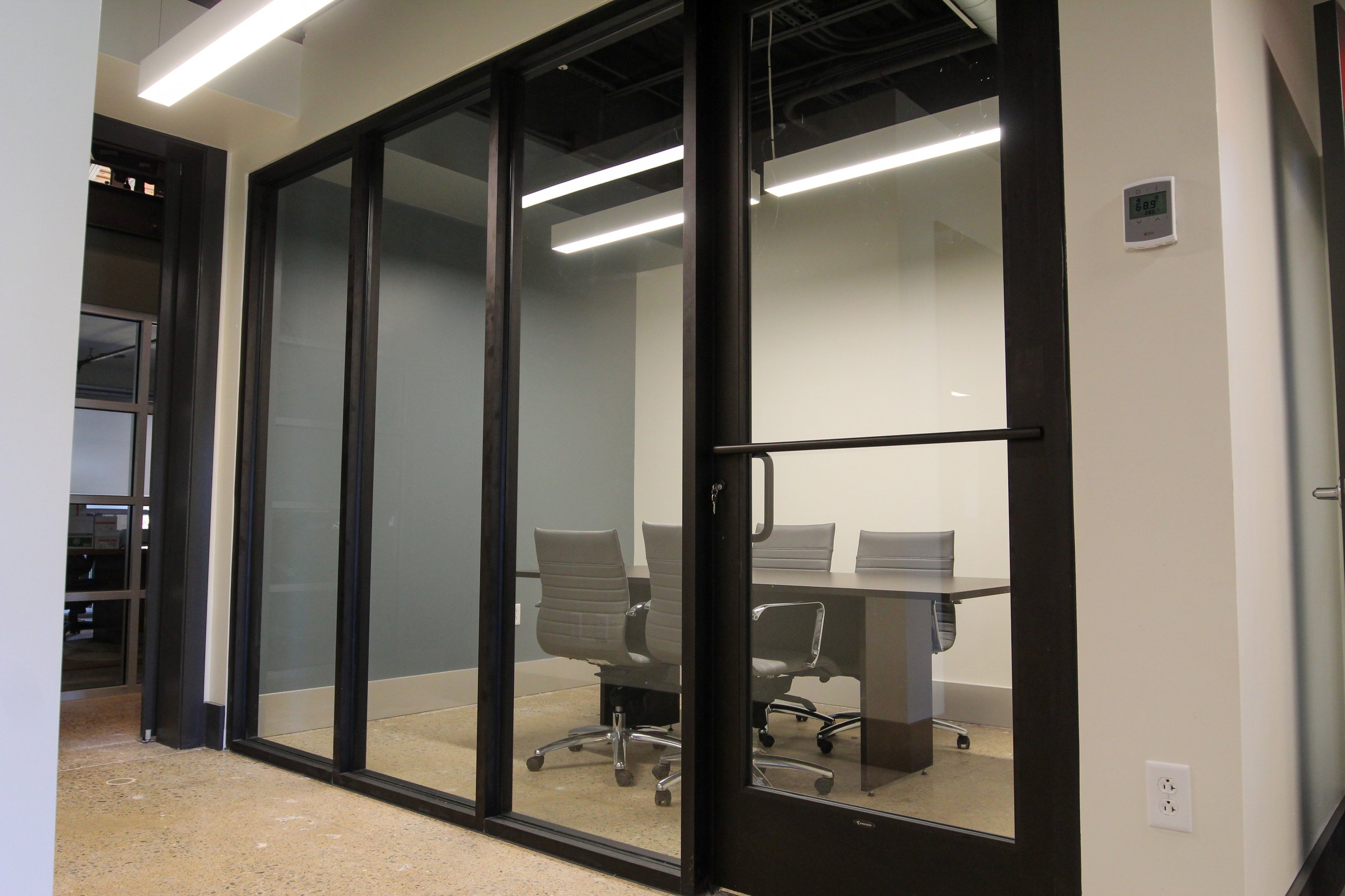 Office conference room with a black-framed glass wall, grey chairs, a table, and a thermostat on the wall.