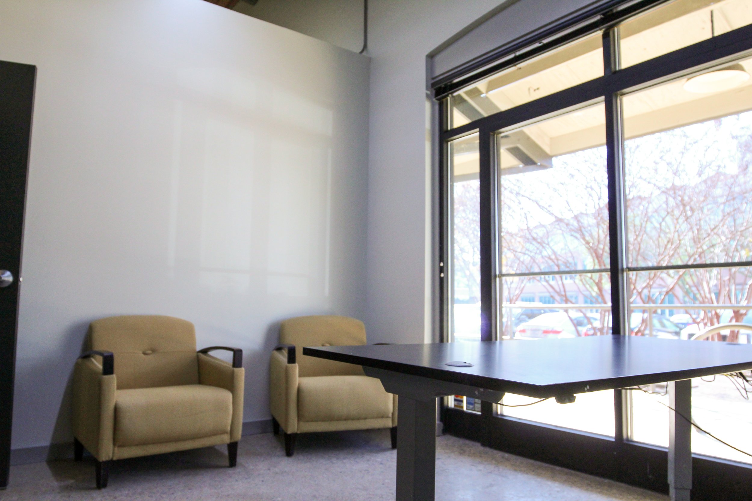Empty office room with two beige chairs, a table, and large window showing parked cars and leafless trees outside.