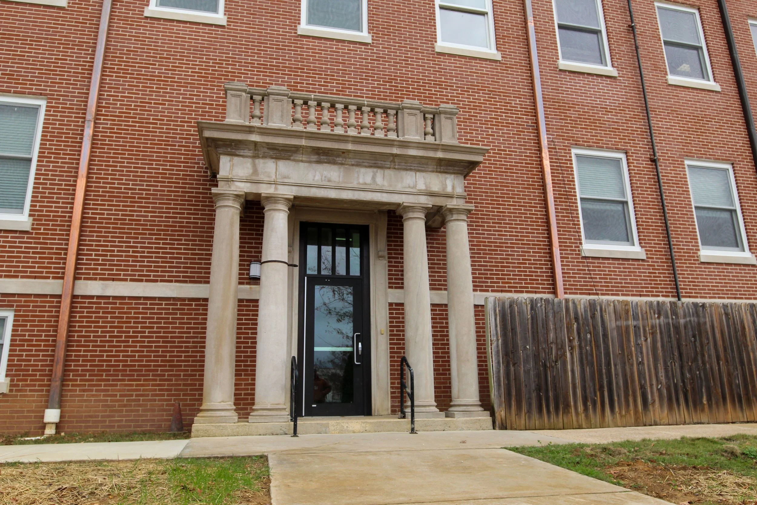 The entrance of a brick apartment building with four windows on the upper floors, a black door with a glass panel, and a stone porch supported by four columns.