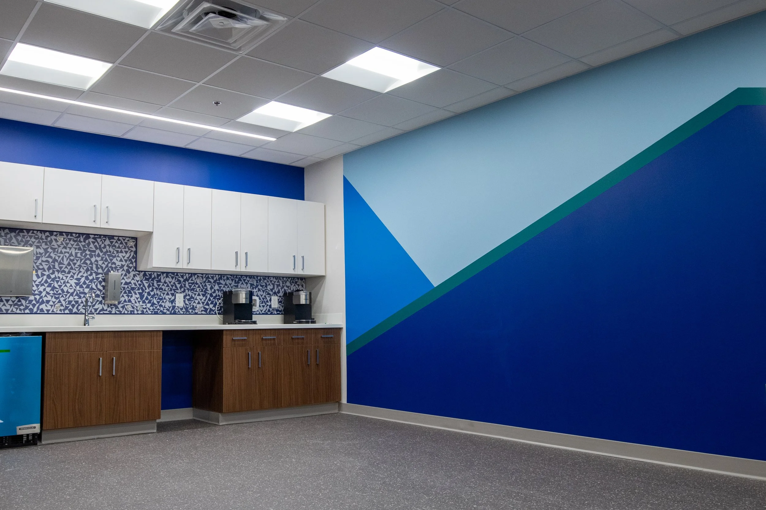 Break room with white and wooden cabinets, coffee machines, a mini fridge, and a geometric blue wall design.