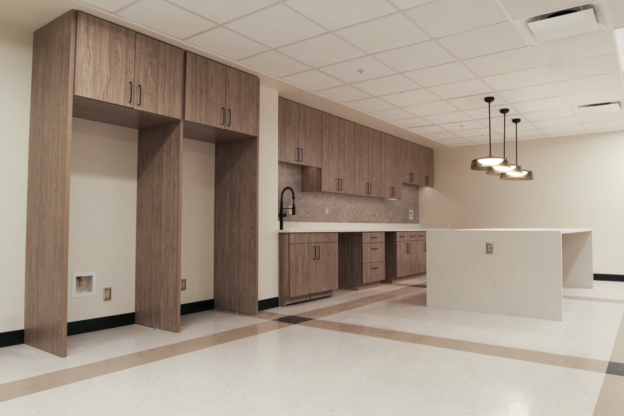 Empty kitchen of Simmons Bank in Athens, TN with wooden cabinets, black faucet, and hanging pendant lights. The floor has a checkered pattern of beige and light brown tiles.