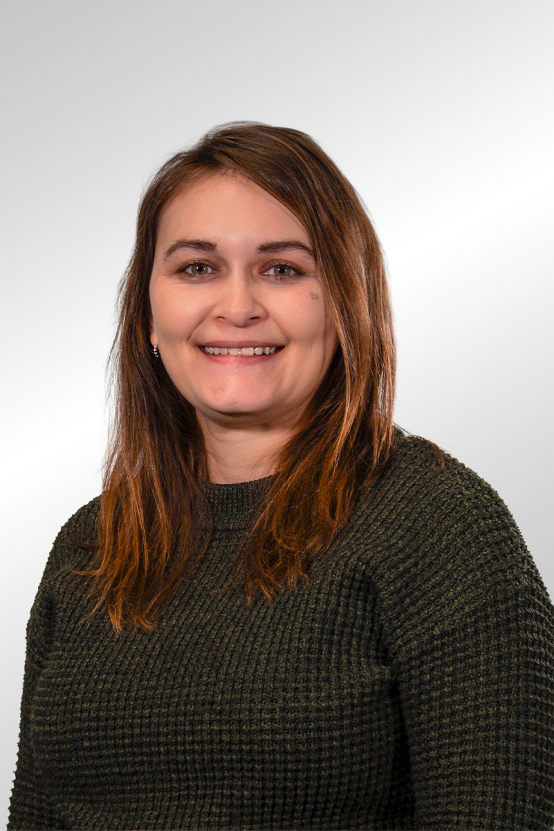 A woman with short dark hair, wearing a black shirt, sitting and looking at the camera with a slight smile, against a dark plain background.