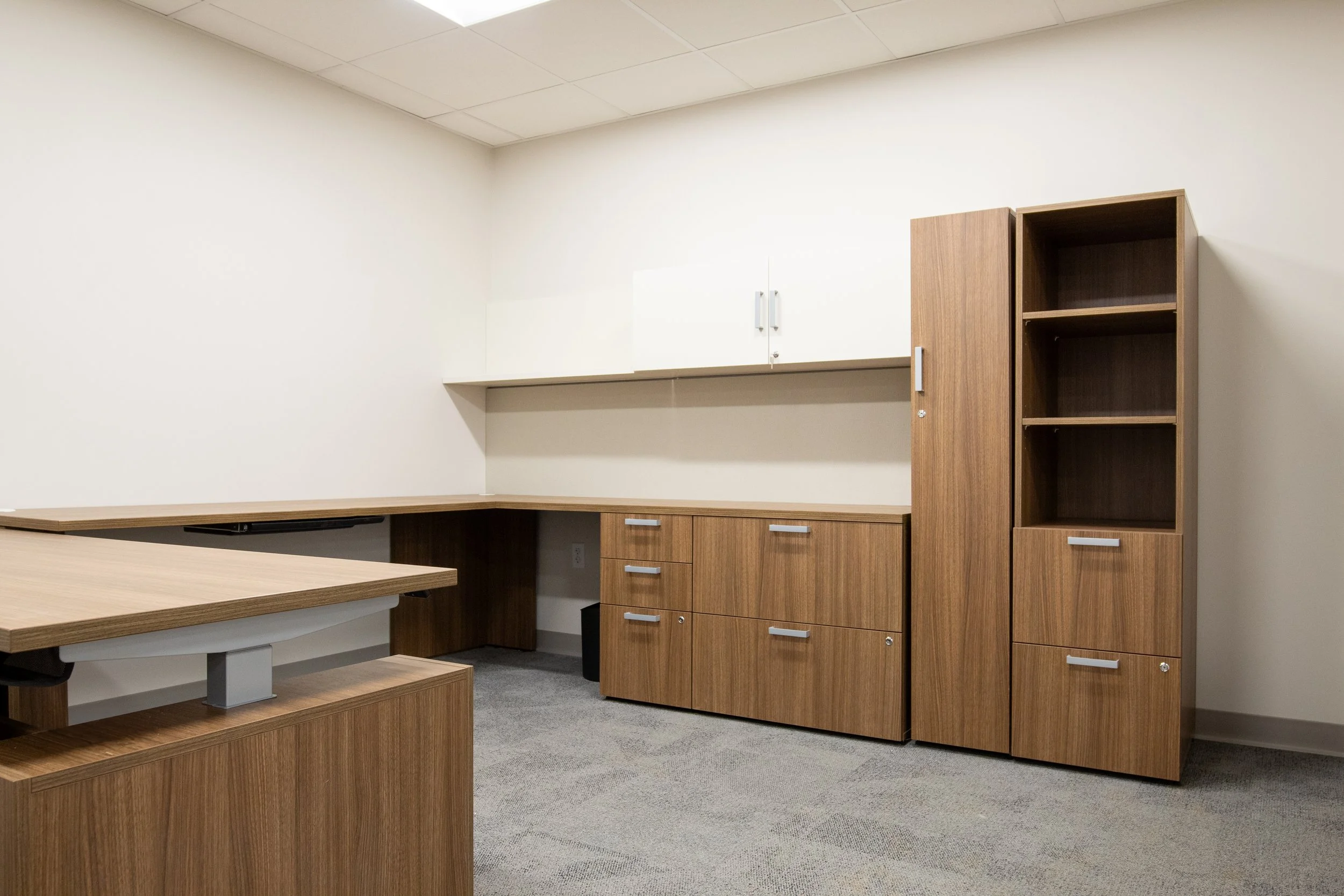 Empty office with wooden desks, cabinets, and shelves against white walls and gray carpet.