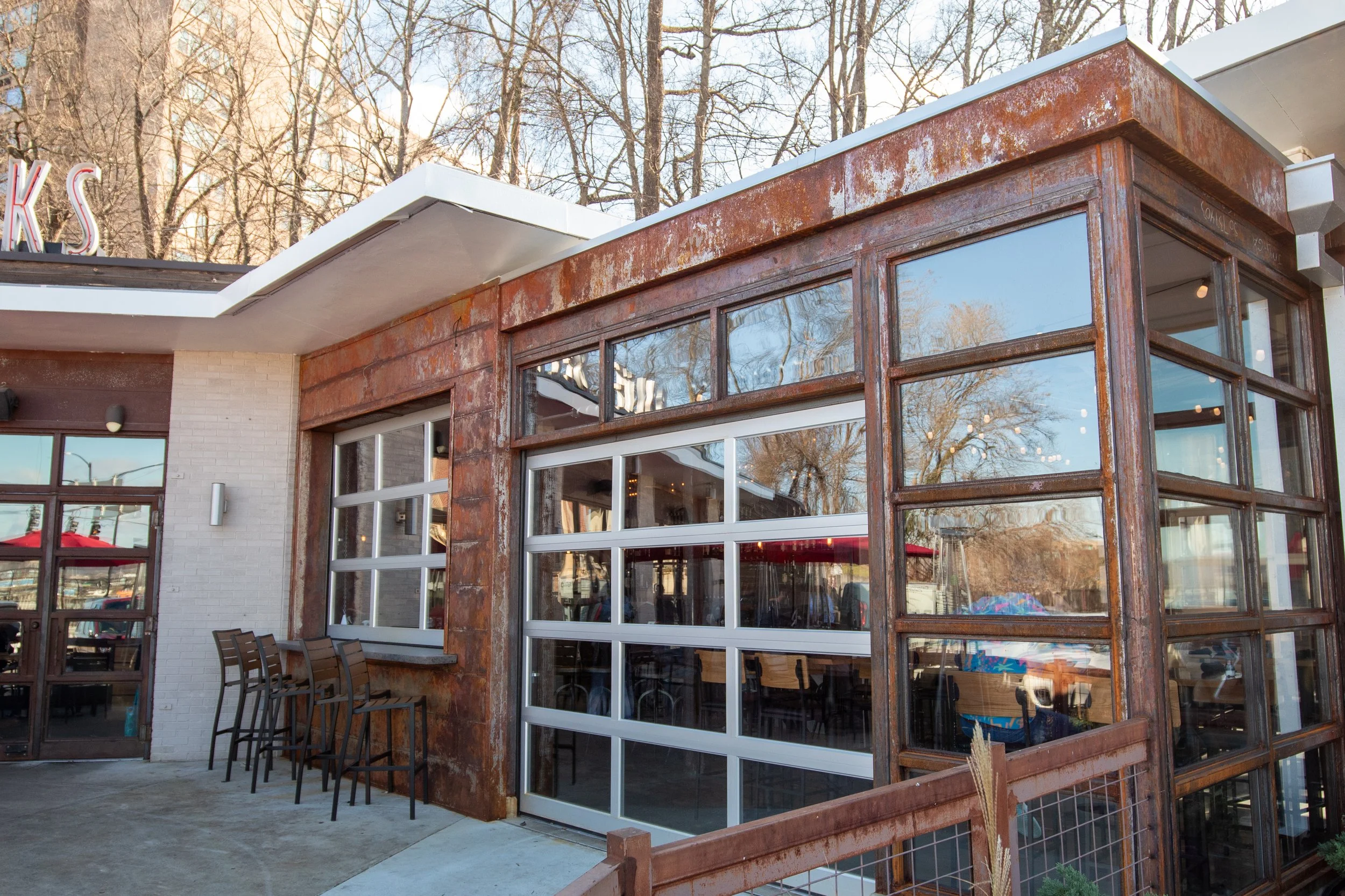 Exterior of a modern restaurant with a rusty metal and glass facade, outdoor seating with chairs, and large windows reflecting trees and the sky.