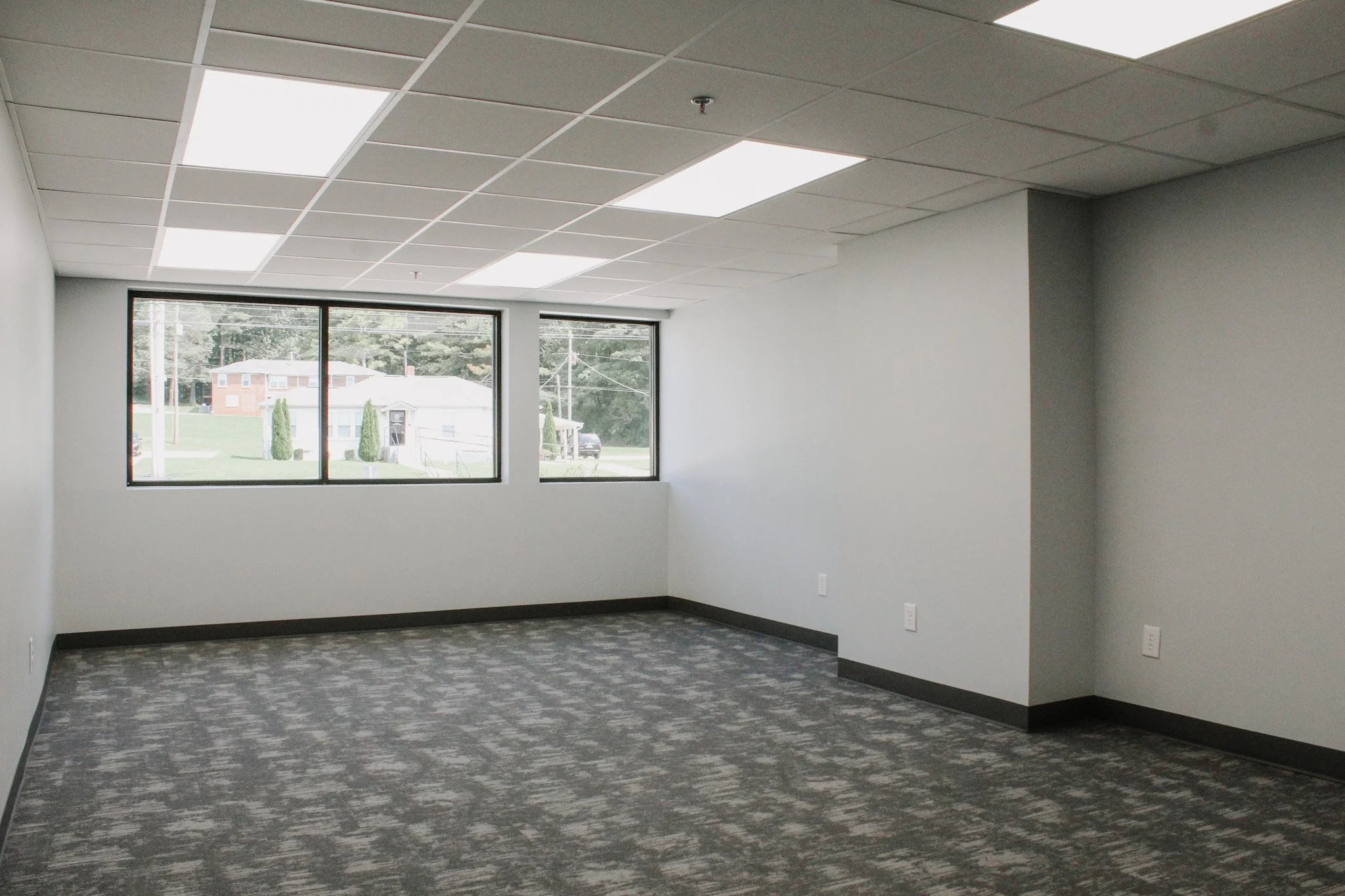 Empty office or conference room with gray walls, carpeted floor, ceiling tiles, and large windows showing a residential neighborhood outside.