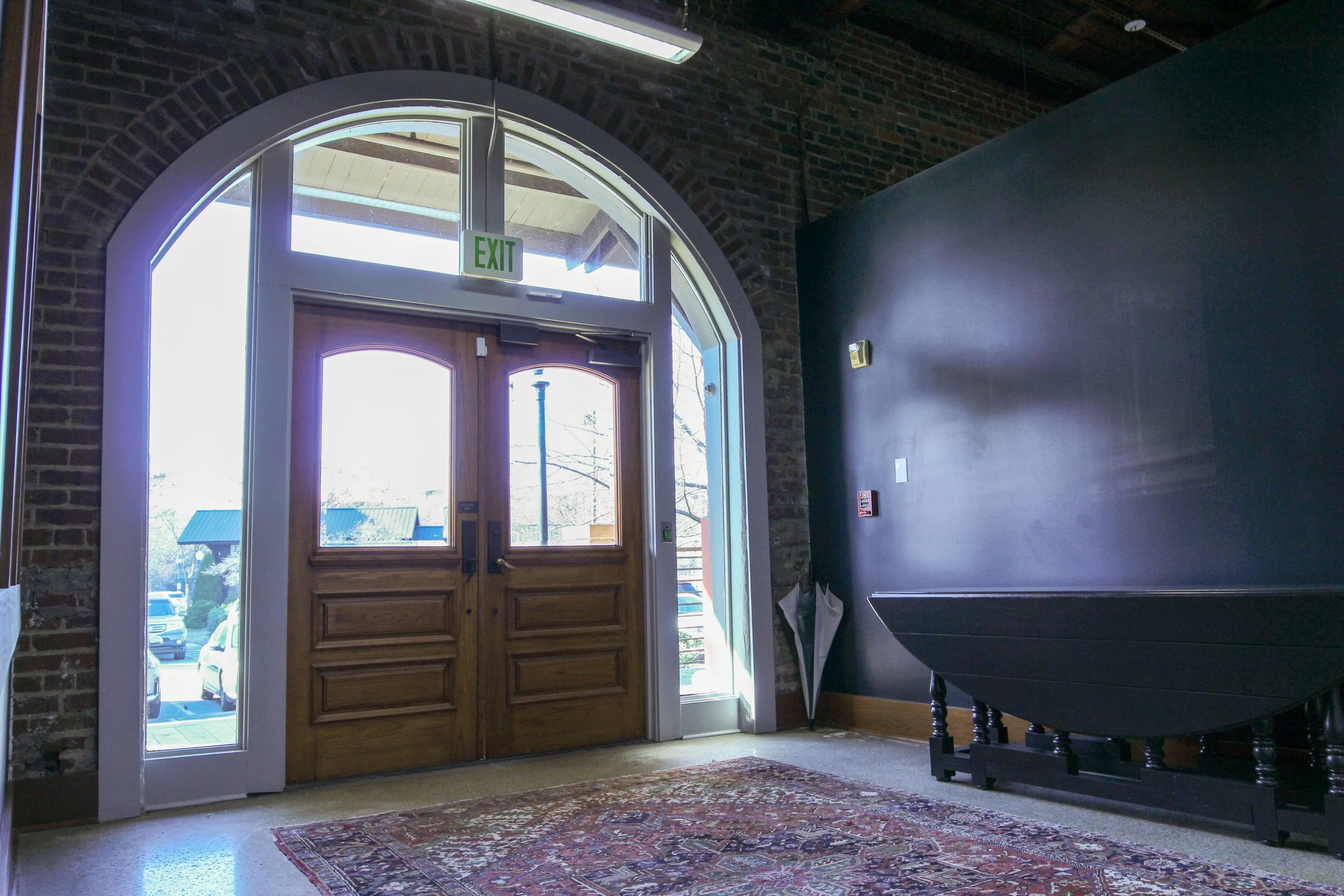 Inside view of a building entrance with wooden double doors, large arched window, brick walls, an 'EXIT' sign above, a carpet on the floor, and a dark-colored piece of furniture against a blue wall.