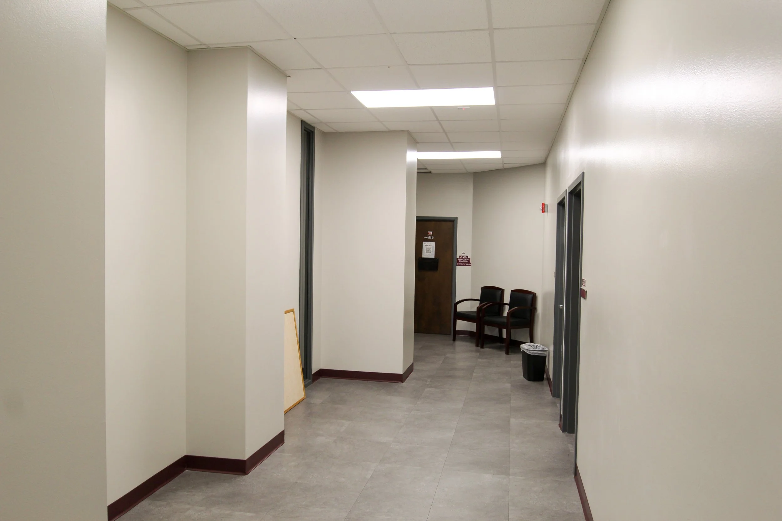 A hallway in a building with beige walls, gray flooring, and a suspended ceiling with fluorescent lights. There are two chairs stacked against the back wall, a trash bin near the right wall, and a closed wooden door at the end of the hallway.