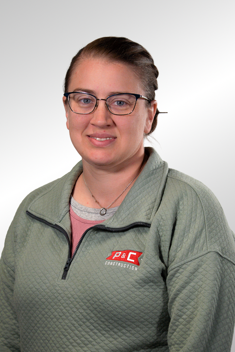A young woman with glasses and brown hair tied back, smiling, wearing a blue and white plaid shirt over a pink shirt, standing against a gray background.