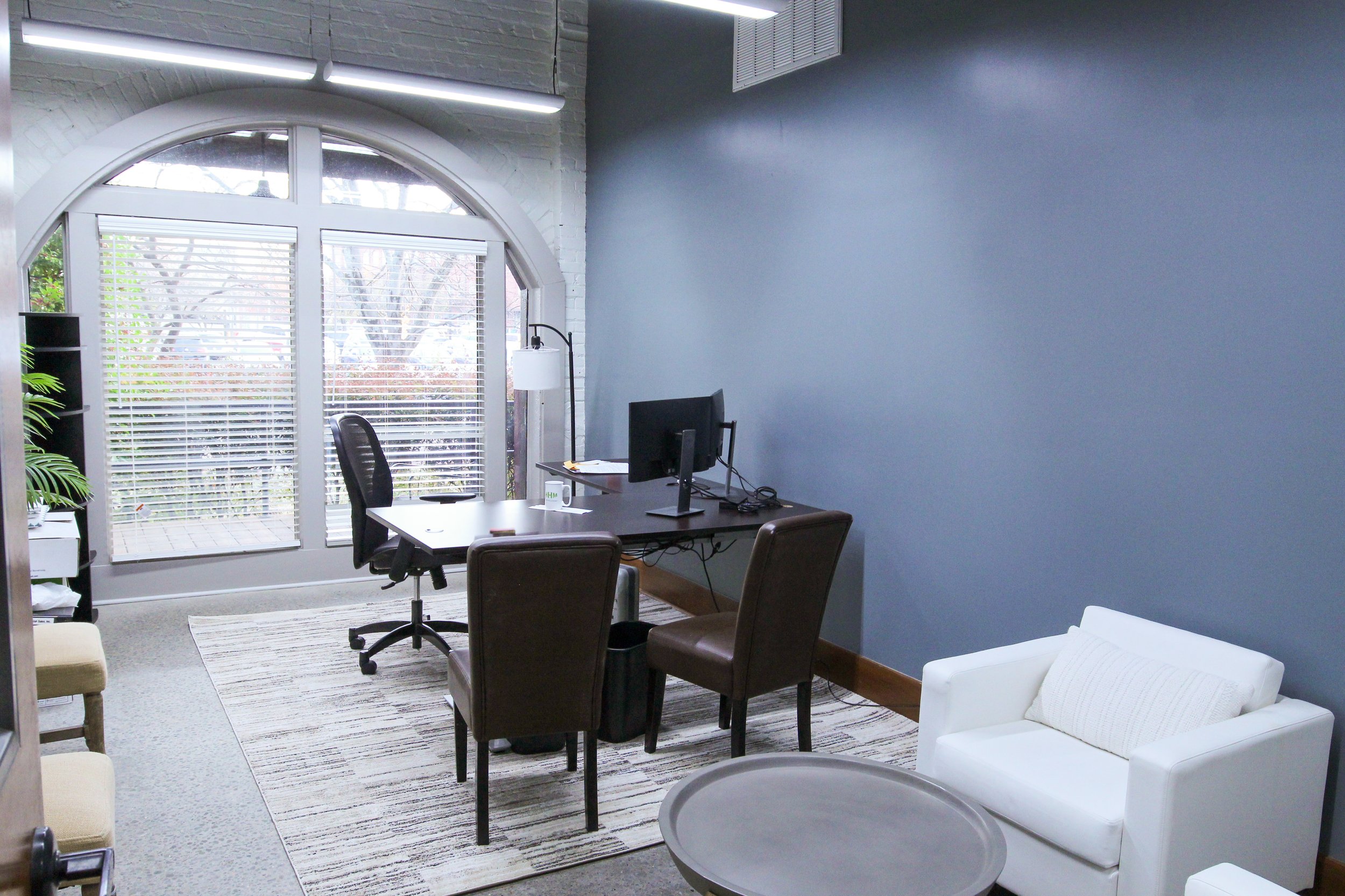An office space with a large arched window with blinds, a desk with a computer monitor, a black office chair, two brown chairs, a white sofa, a small round table, a standing lamp, and a gray wall.