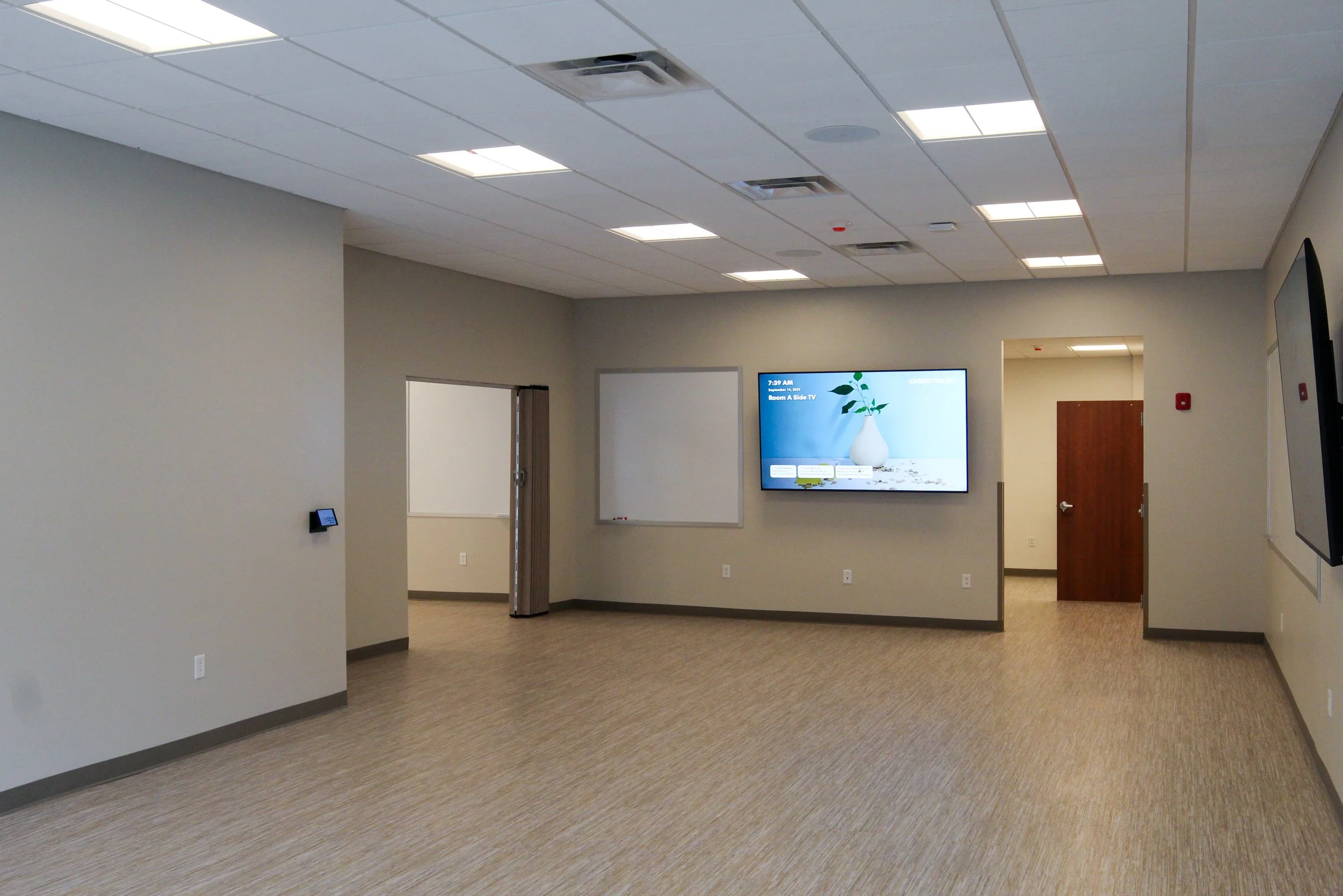 Empty conference or meeting room with walls painted light gray, beige carpet, ceiling lights, wall-mounted digital display, and whiteboards.