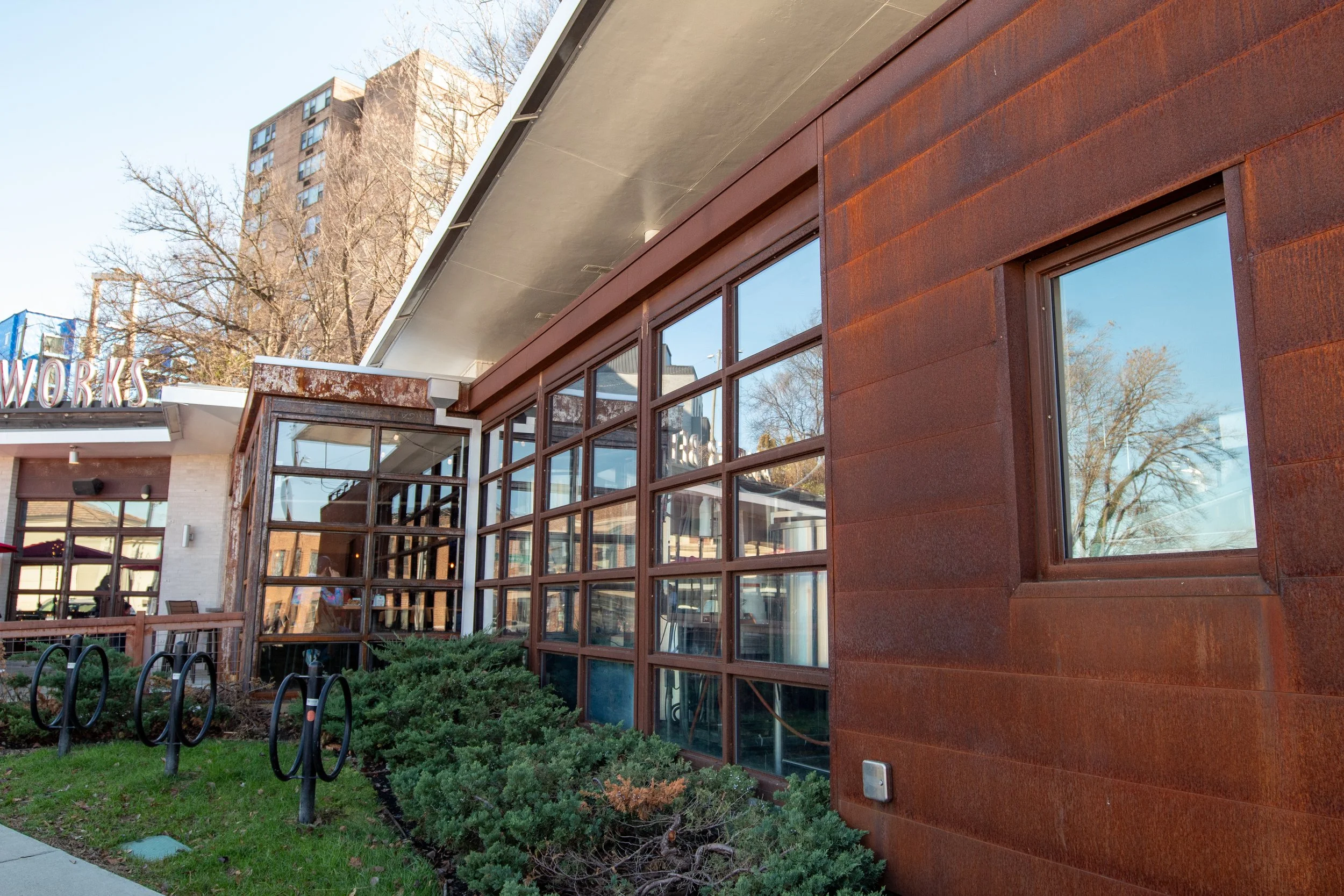 Exterior view of a modern building with large glass windows and rust-colored siding, surrounded by greenery and bike racks.