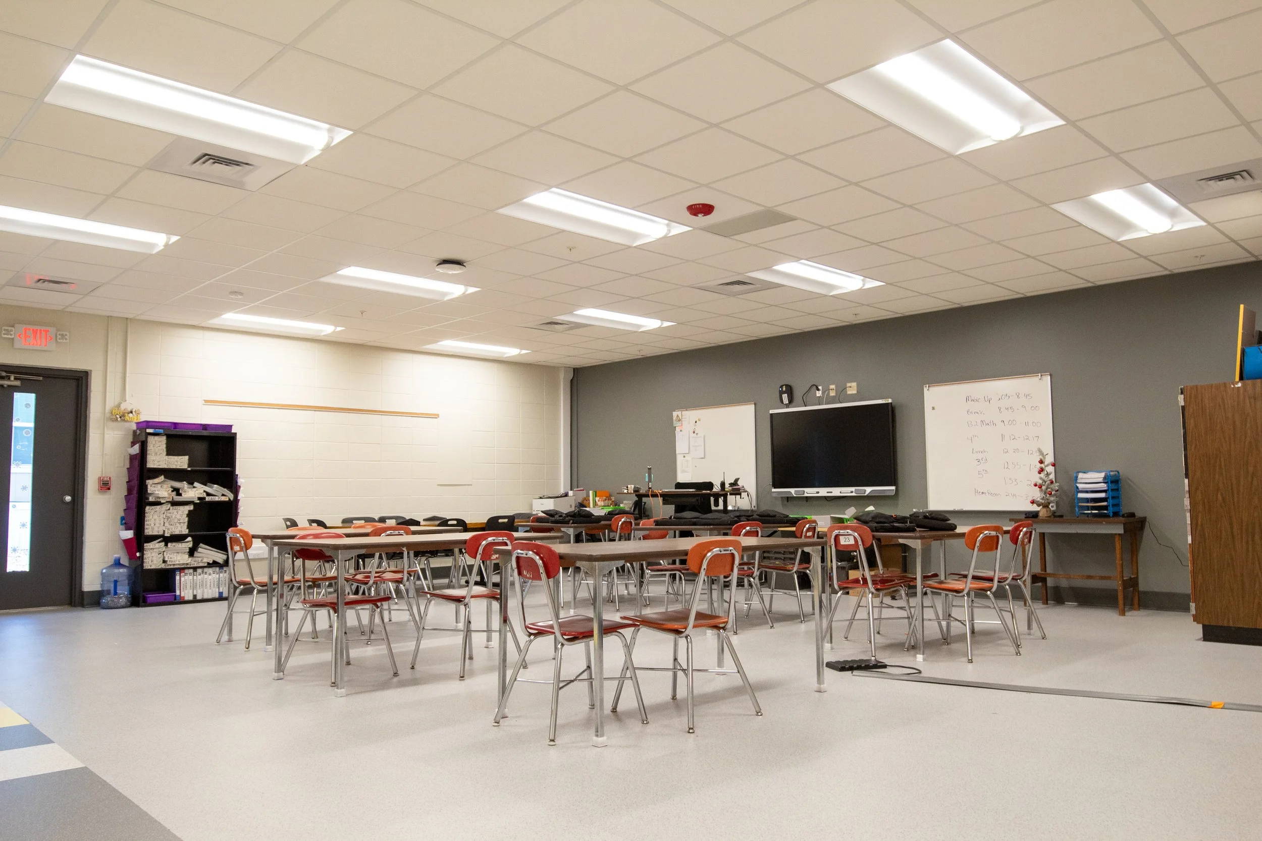 Empty classroom with tables and chairs, whiteboard, TV, and storage shelves.