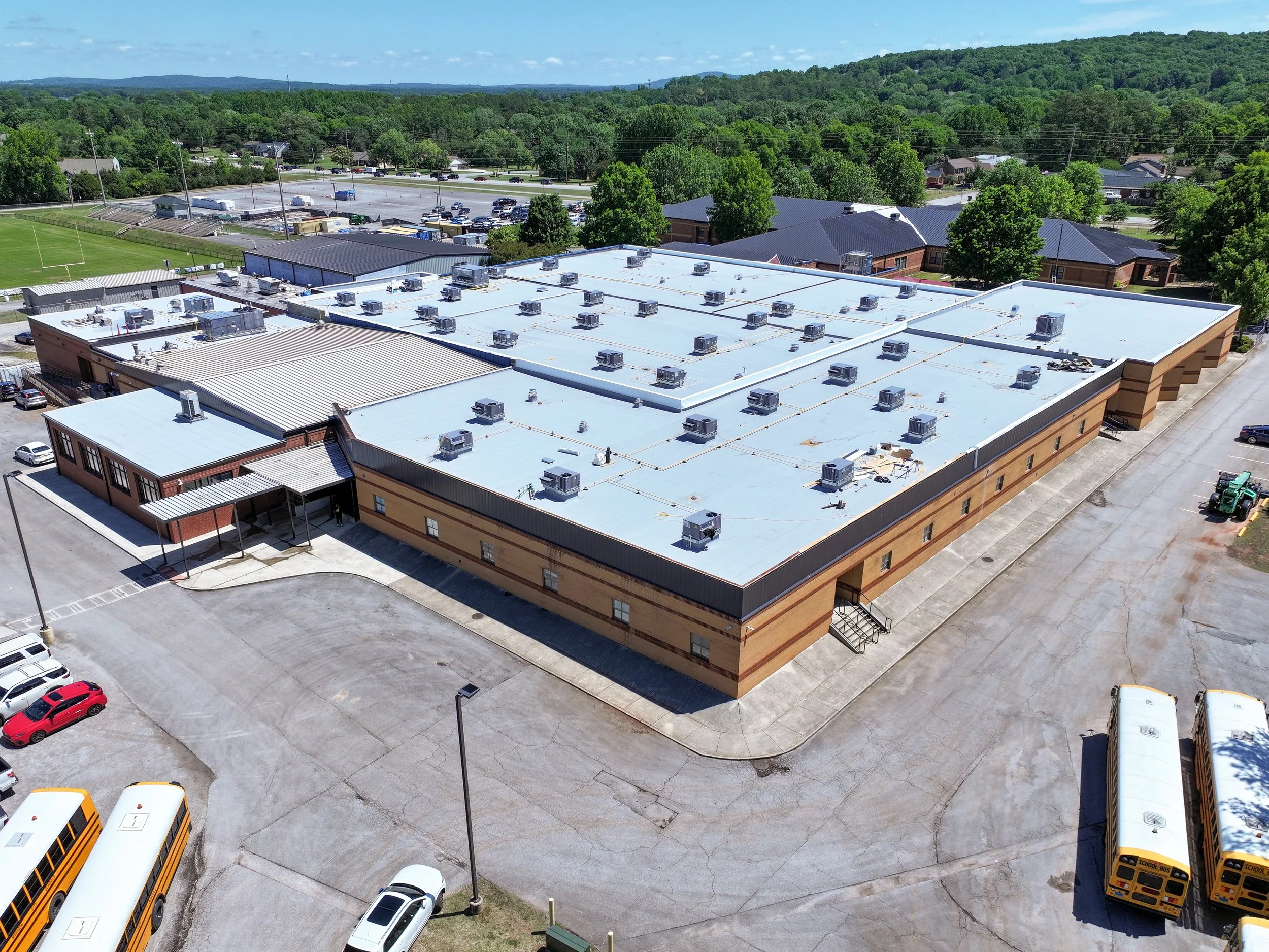 Aerial view of a school building with a white roof and multiple air conditioning units, surrounded by parking lot and nearby fields with green trees and hills in the background. Remodeled by P&C Construction, Inc. - Chattanooga, Tn.